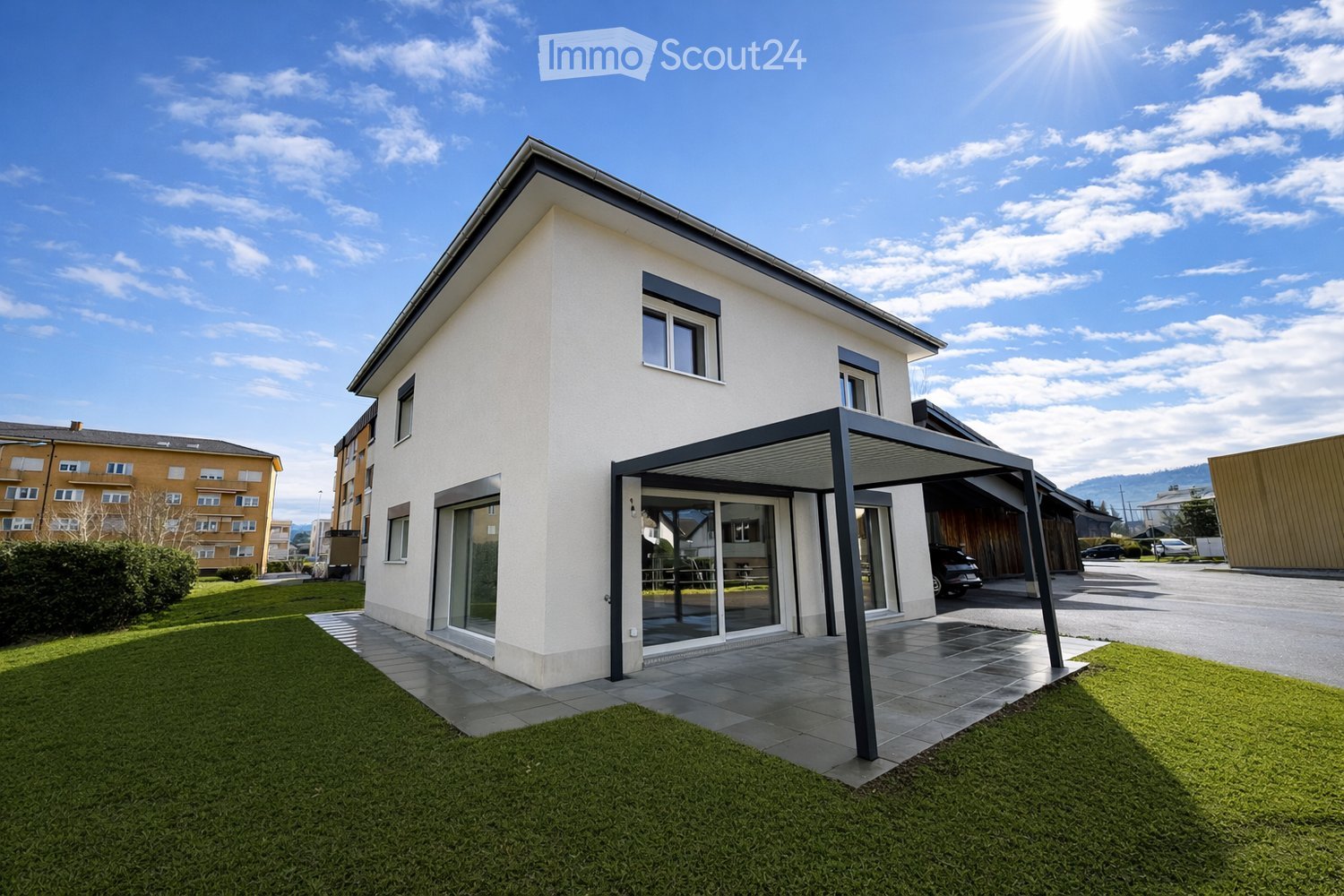 Modern two-story house with white exterior, large windows, and a covered patio area. The house features a well-maintained lawn and is situated in a residential area with neighboring buildings visible in the background.