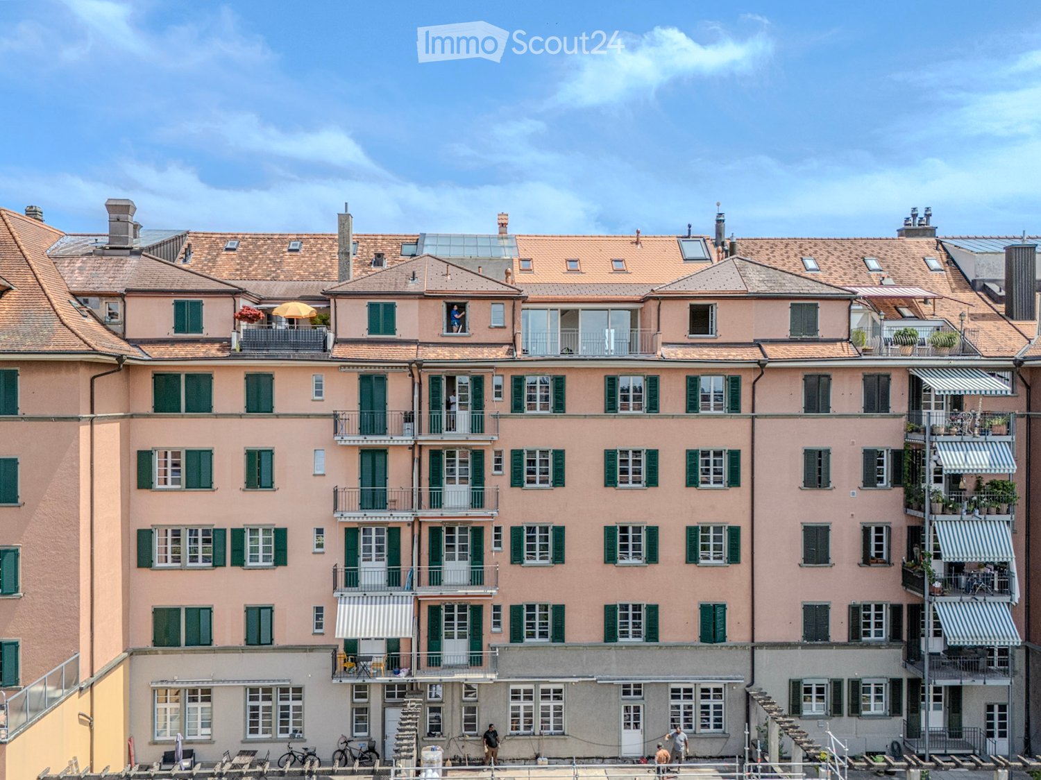 Multi-story residential building with pink facades, green shutters, balconies, and a tiled roof.