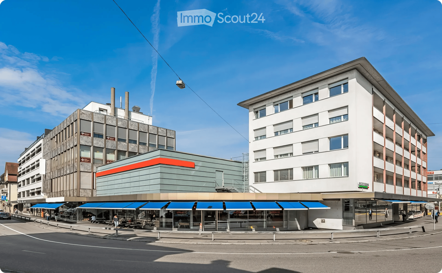 A multi-story building with a modern facade, featuring a mix of glass and concrete elements, with a restaurant area and seating under blue awnings.