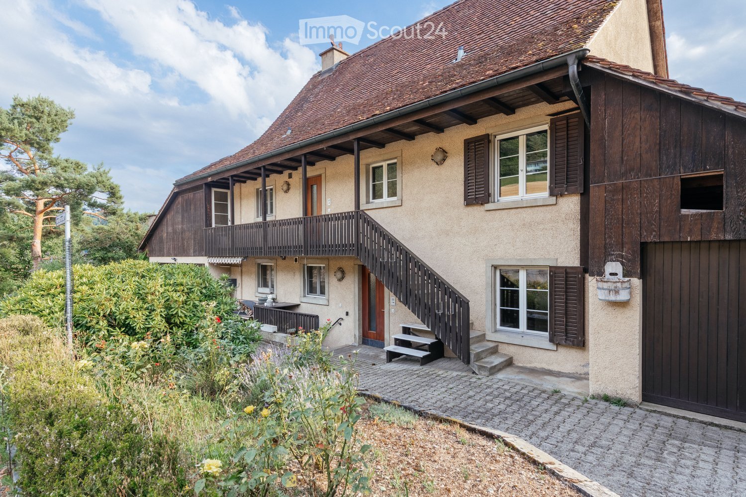 Two-story house with a tiled roof, wooden balcony, and stone facade. Features include multiple windows with shutters, a wooden staircase leading to the entrance, and a small garden area with plants and a bench.