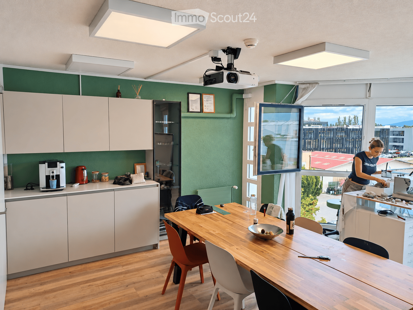 Modern kitchen with light-colored cabinets, a wooden dining table with chairs, a coffee machine, and a projector mounted on the ceiling. The room features large windows with a view of the outdoor area.