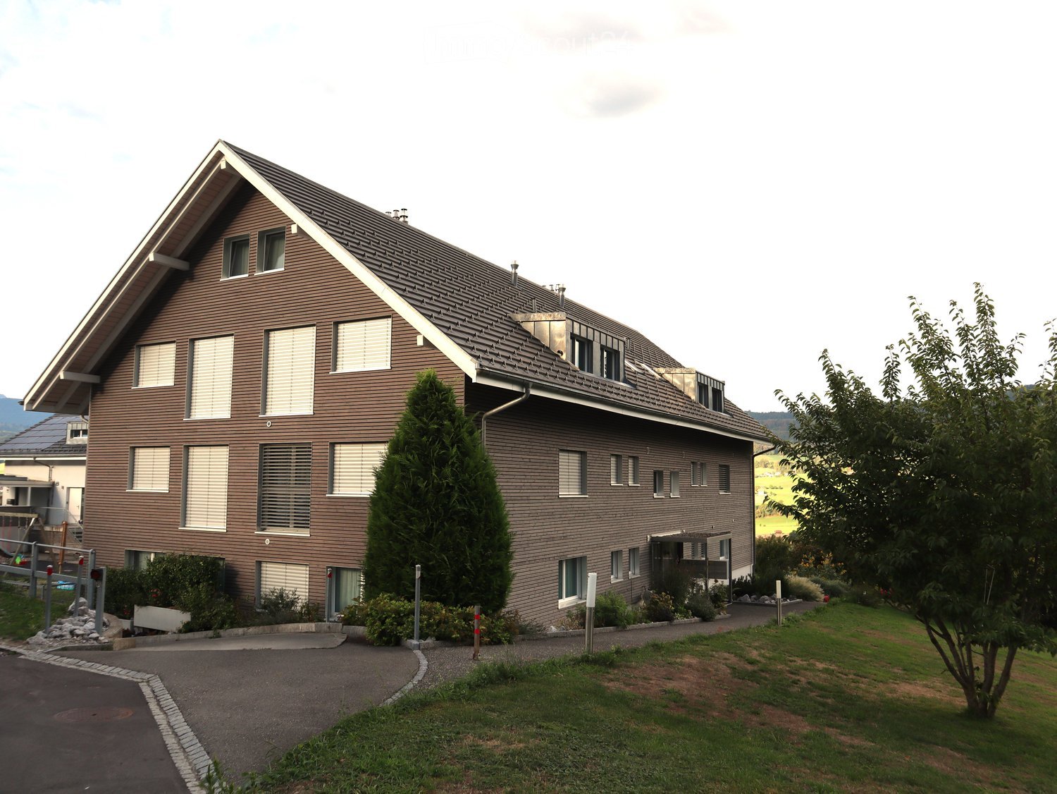 Two-story house with brown siding, multiple windows, and a sloped roof. The house is surrounded by a well-maintained lawn, trees, and a driveway.