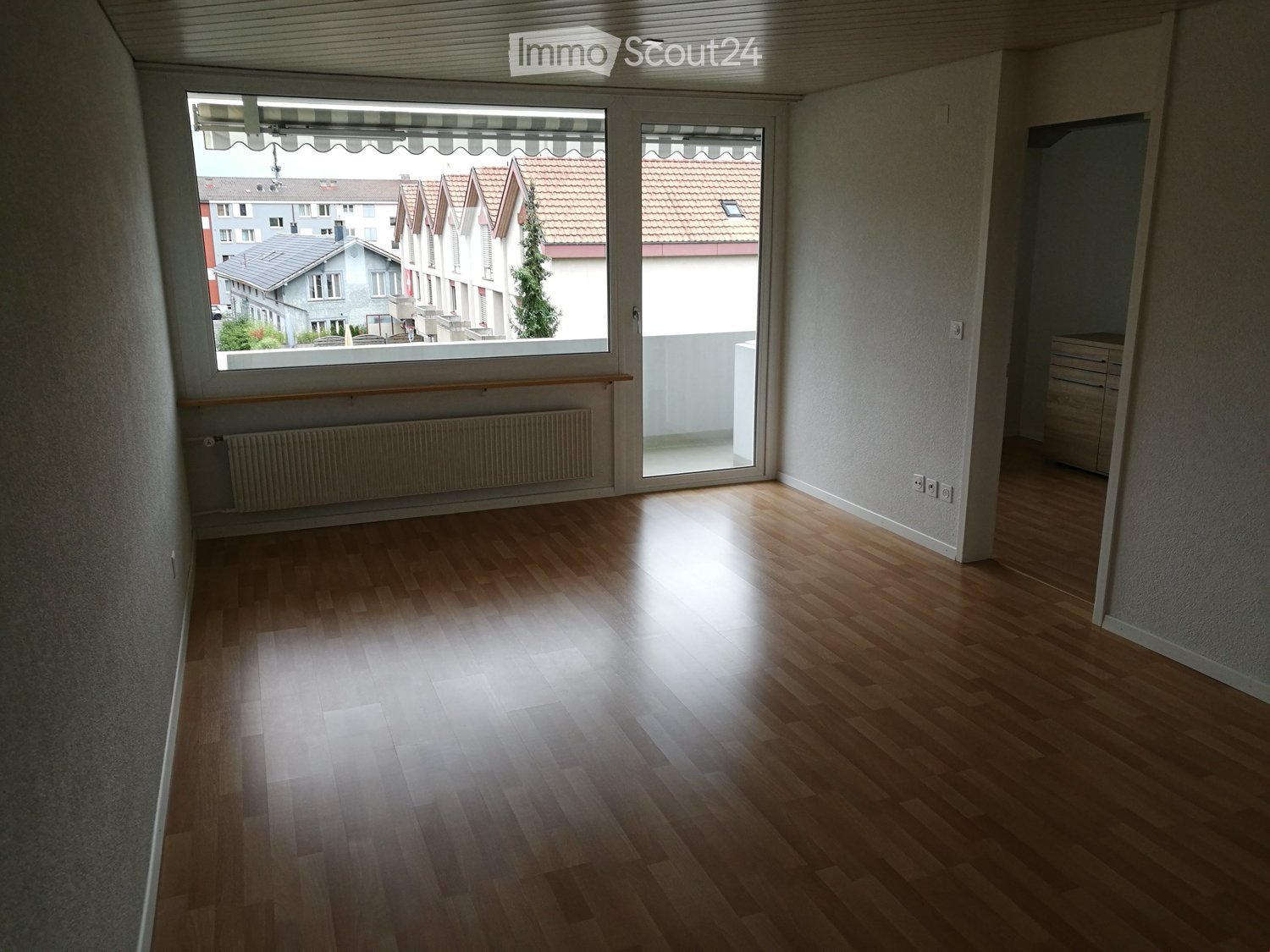 Empty room with wooden flooring, large window with view of neighboring houses, radiator below window, and a sliding glass door leading to a balcony.