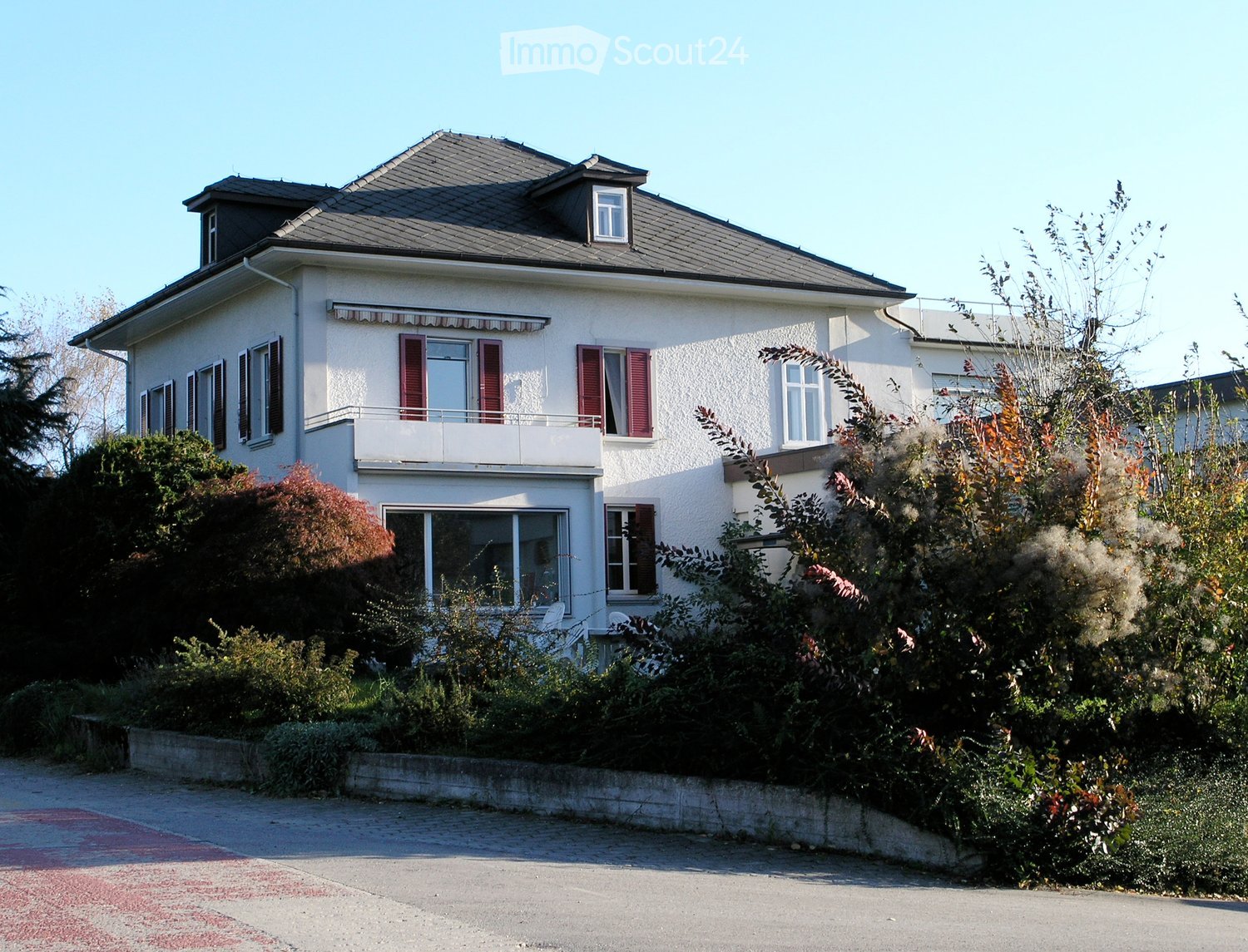 Two-story house with white exterior, red shutters, and a balcony on the upper level. The house is surrounded by greenery and has a driveway leading to it.