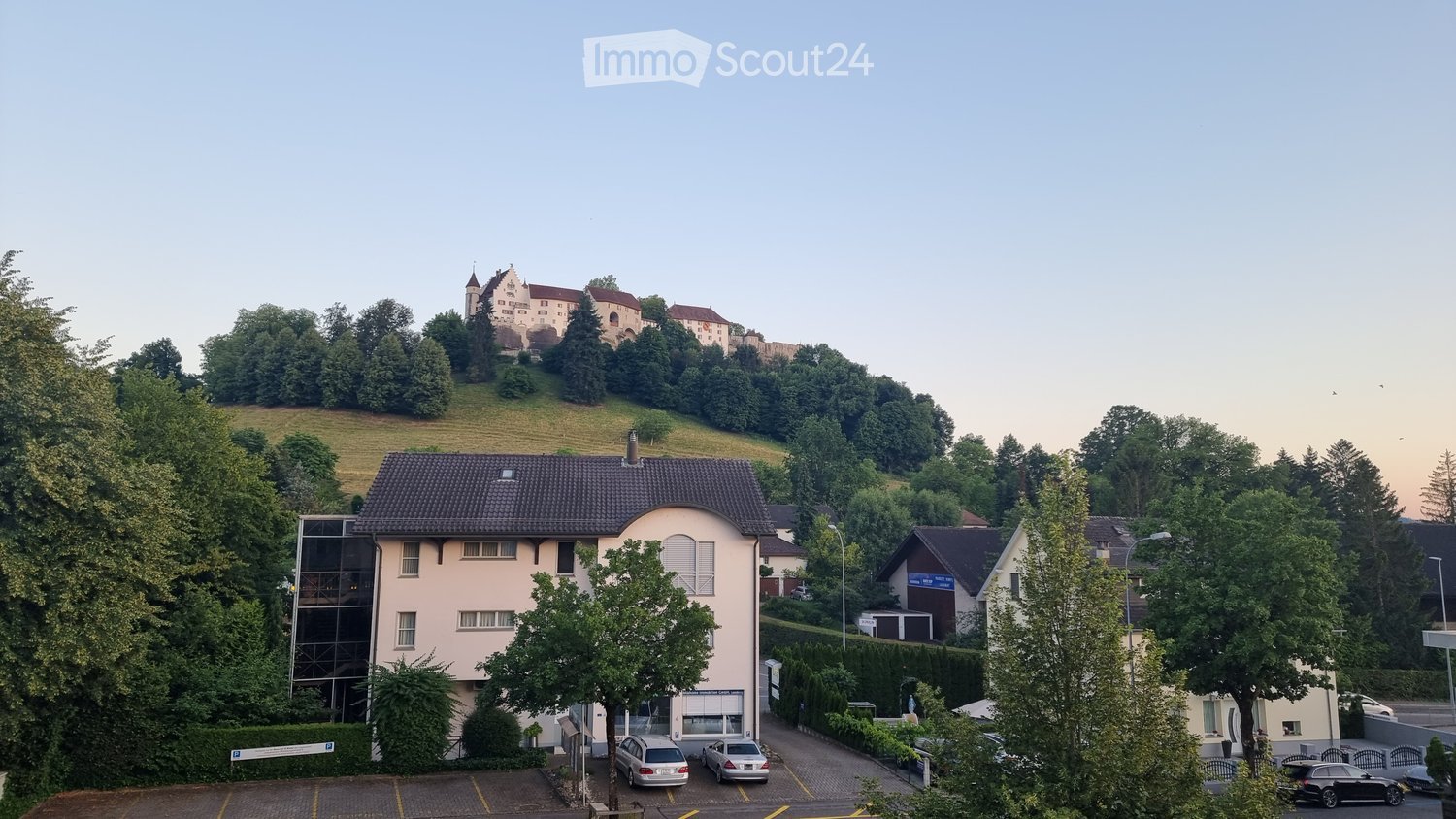 A modern two-story building with a tiled roof, surrounded by trees and a parking lot, with a historic castle visible on a hill in the background.
