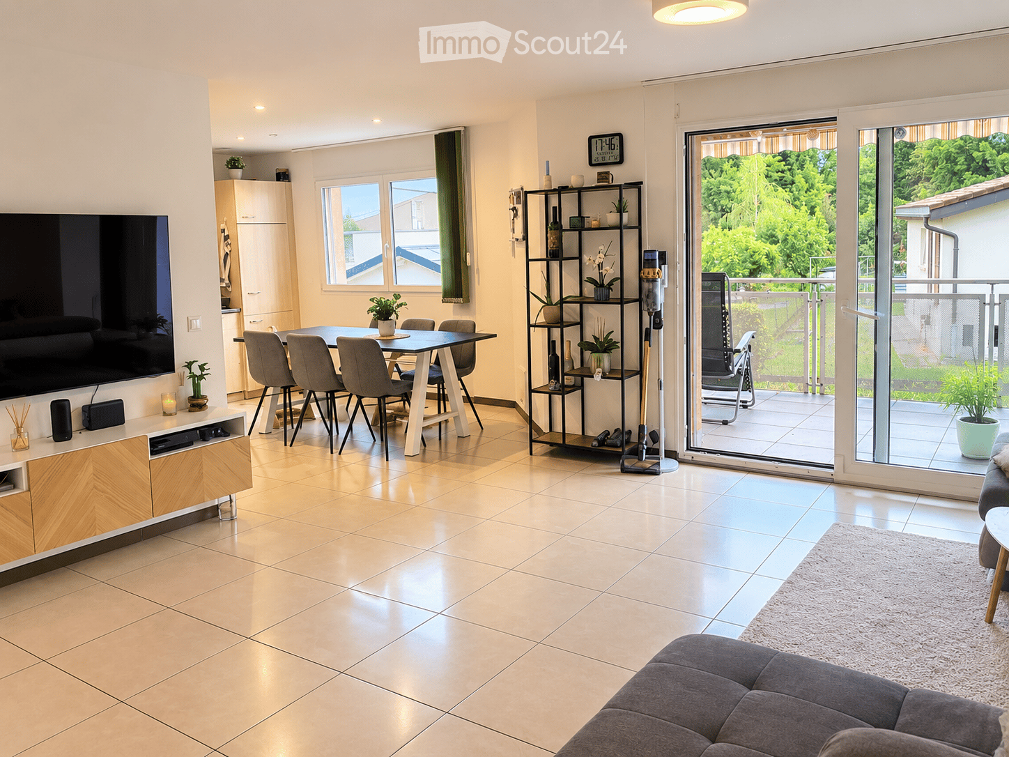 Modern living and dining area with a grey sofa, coffee table, dining table with chairs, shelves, and a large TV screen. The space features large windows and a sliding glass door leading to a balcony.