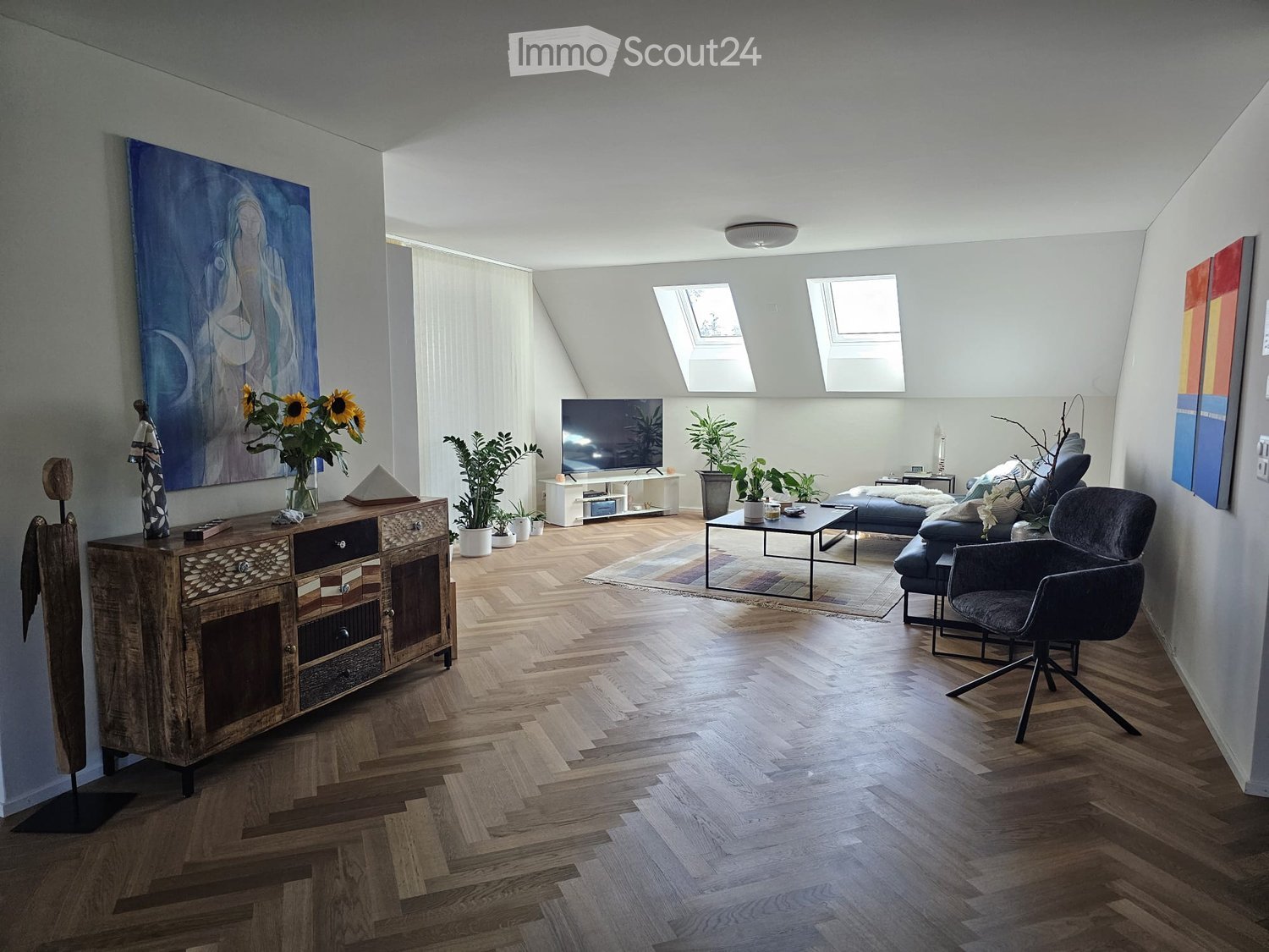 Modern living room with wooden herringbone flooring, large windows, skylights, a sofa, coffee table, armchair, TV, cabinet, and decorative plants.