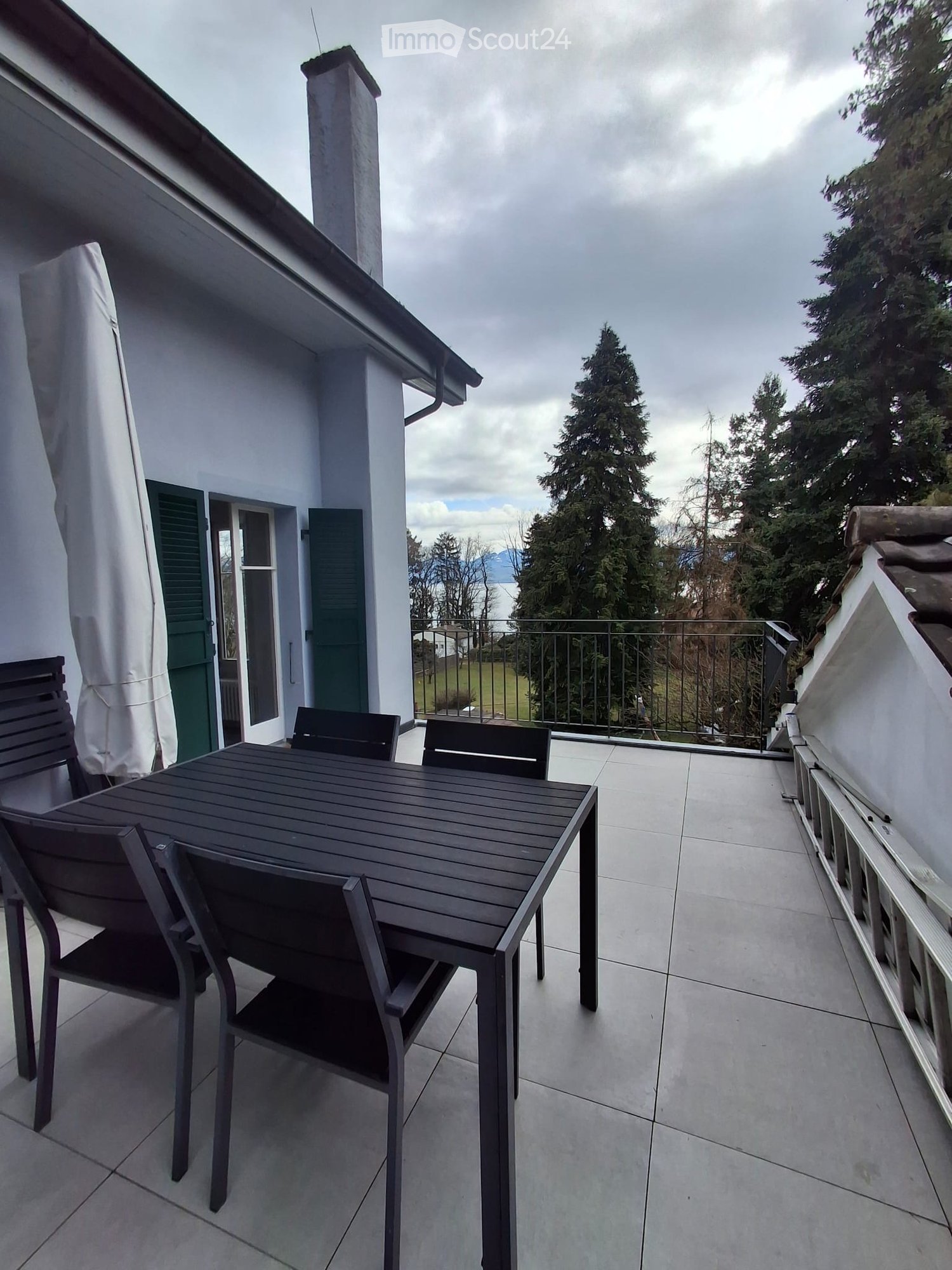 Balcony with a wooden table and four chairs, overlooking a scenic view of trees and a body of water.