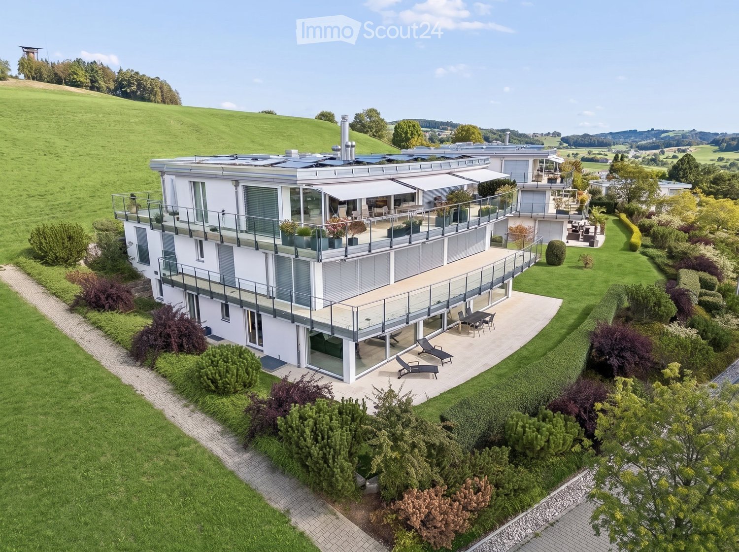 Modern two-story house with large balconies, surrounded by a well-maintained garden and lawn area.