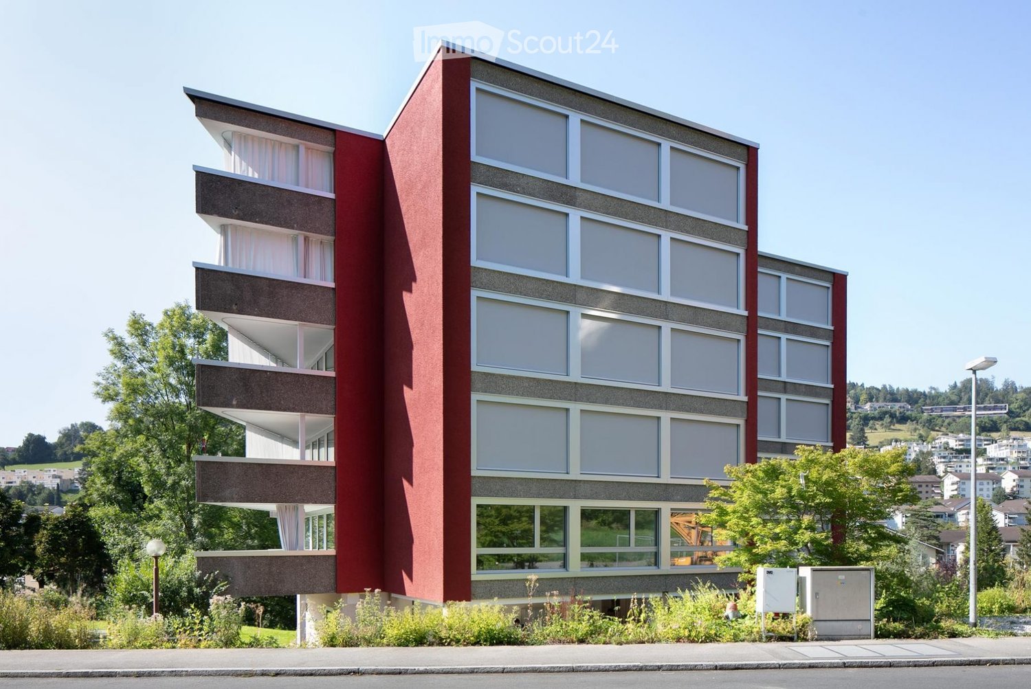 Modern multi-story building with red and gray facade, large windows, and balconies. The building is surrounded by greenery and trees, with a clear blue sky in the background.