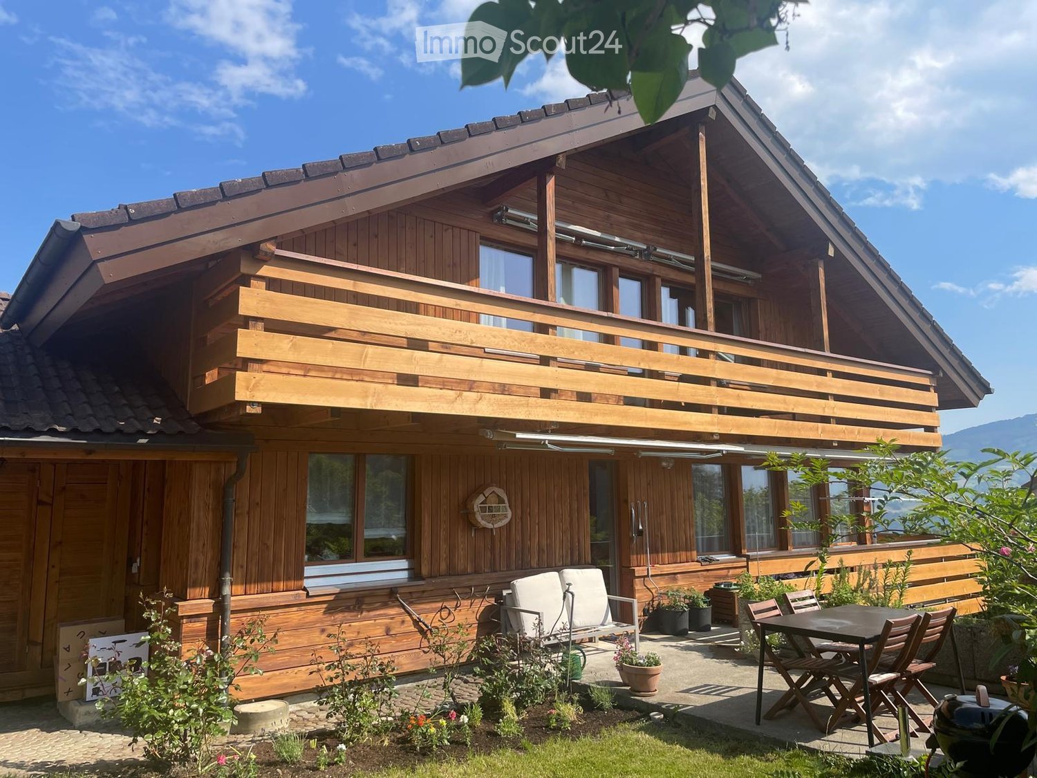 Two-story wooden house with a balcony, large windows, and a well-maintained garden area with outdoor furniture.