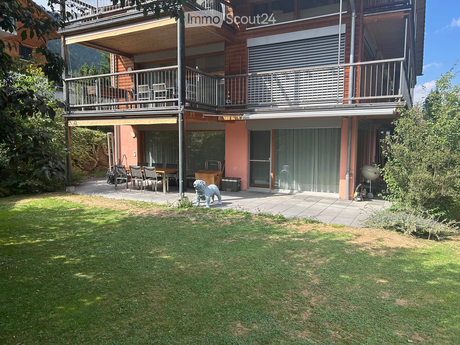 A well-maintained garden with a lawn, patio area, outdoor dining set, and a large wooden house with balconies and large windows.