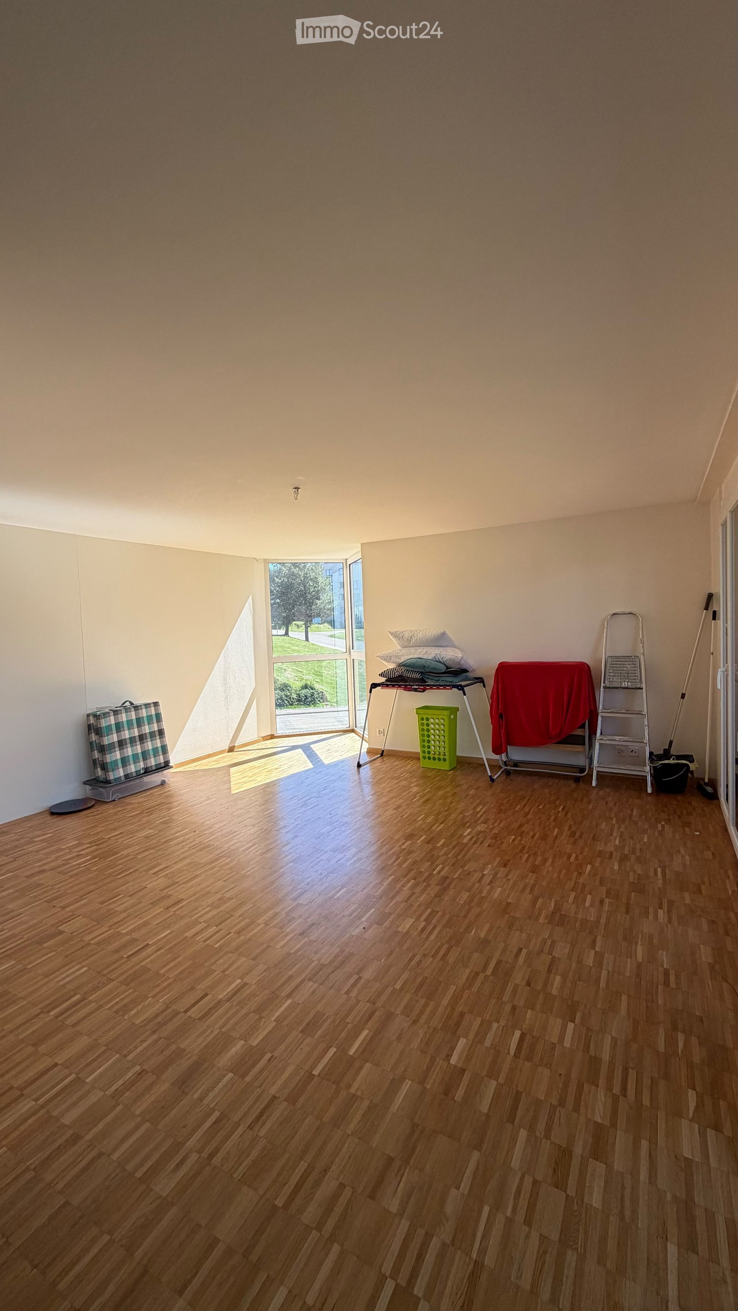 Empty room with wooden flooring, large window, and some storage items including a stool, ladder, and suitcase.