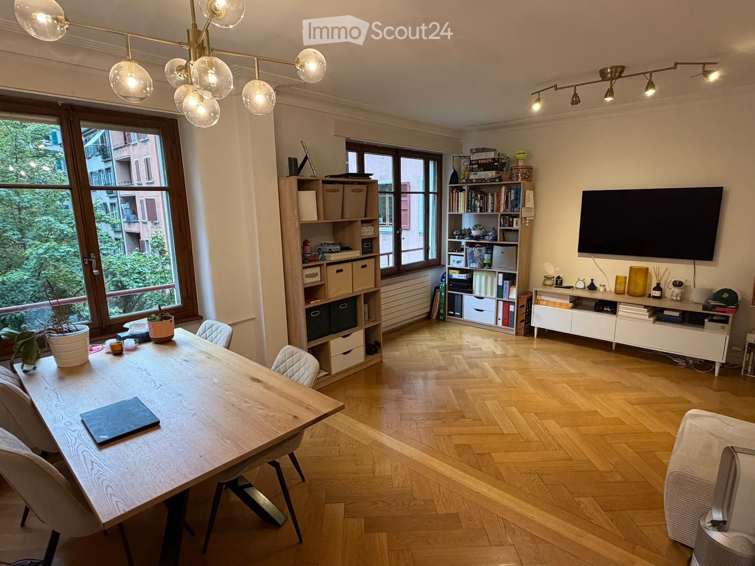 Bright, modern room with wooden flooring, large windows, a dining table with chairs, a laptop, a bookshelf, a TV, and decorative items.