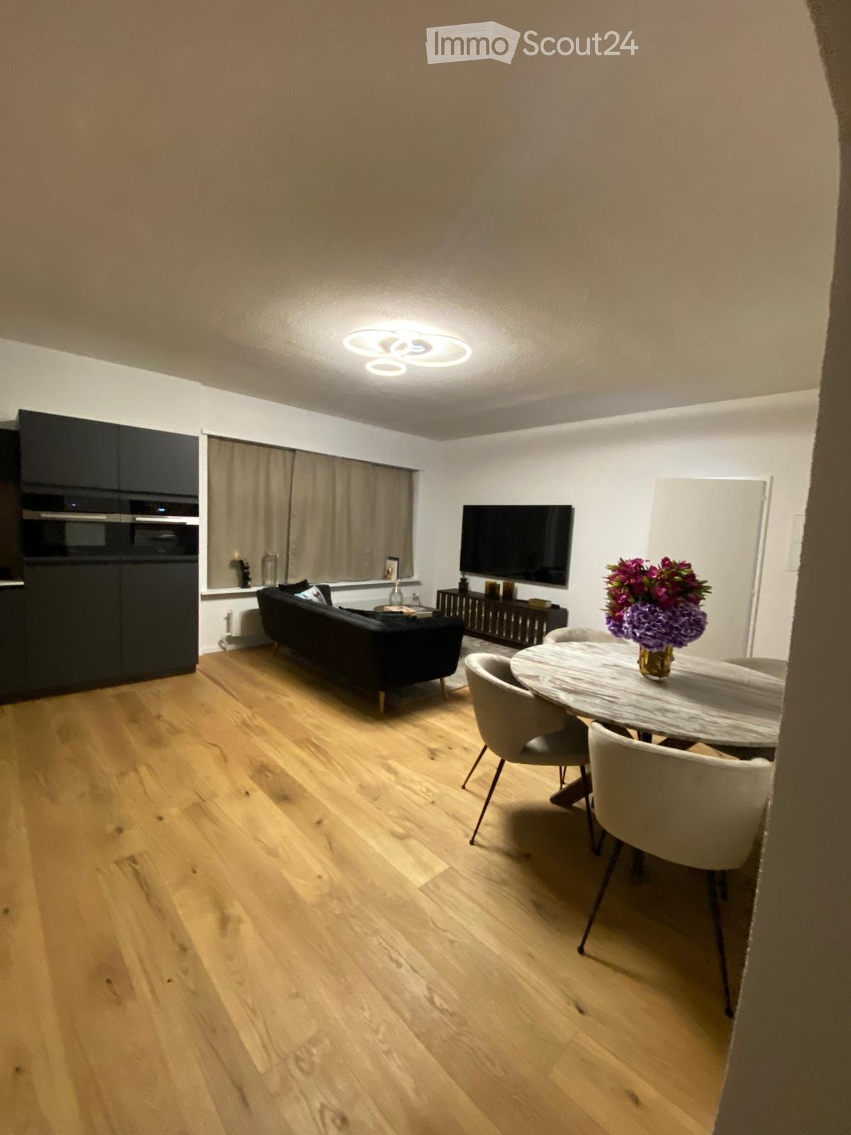 Modern living and dining area with light wooden flooring, a black sofa, a round dining table with chairs, a television mounted on the wall, and a flower vase on the table.