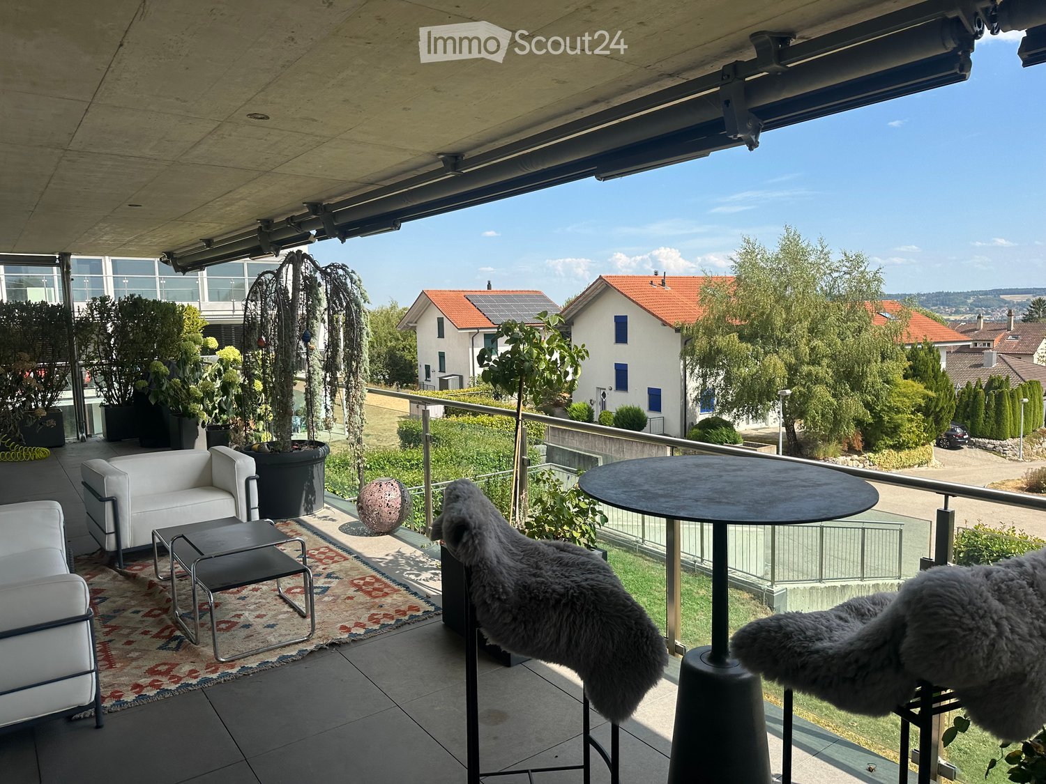 Balcony with modern furniture including a white sofa, a round table, a stool, a rug, and two furry chairs. Potted plants and a scenic view of the neighborhood are visible.