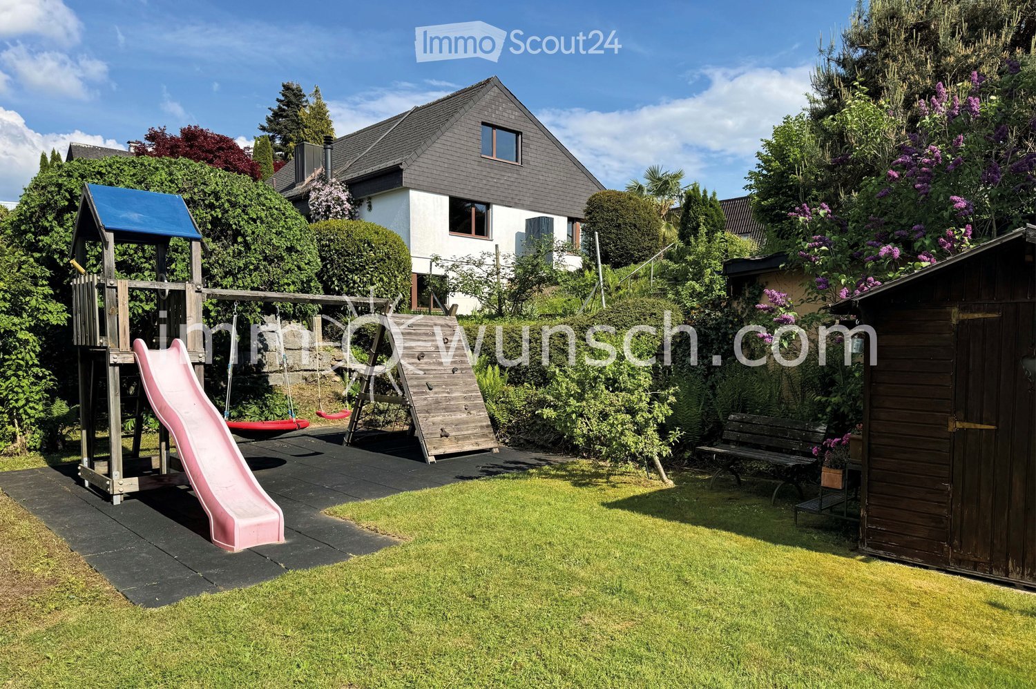 Well-maintained garden with a wooden play structure featuring a slide, swings, and climbing wall, surrounded by lush greenery and a wooden shed.