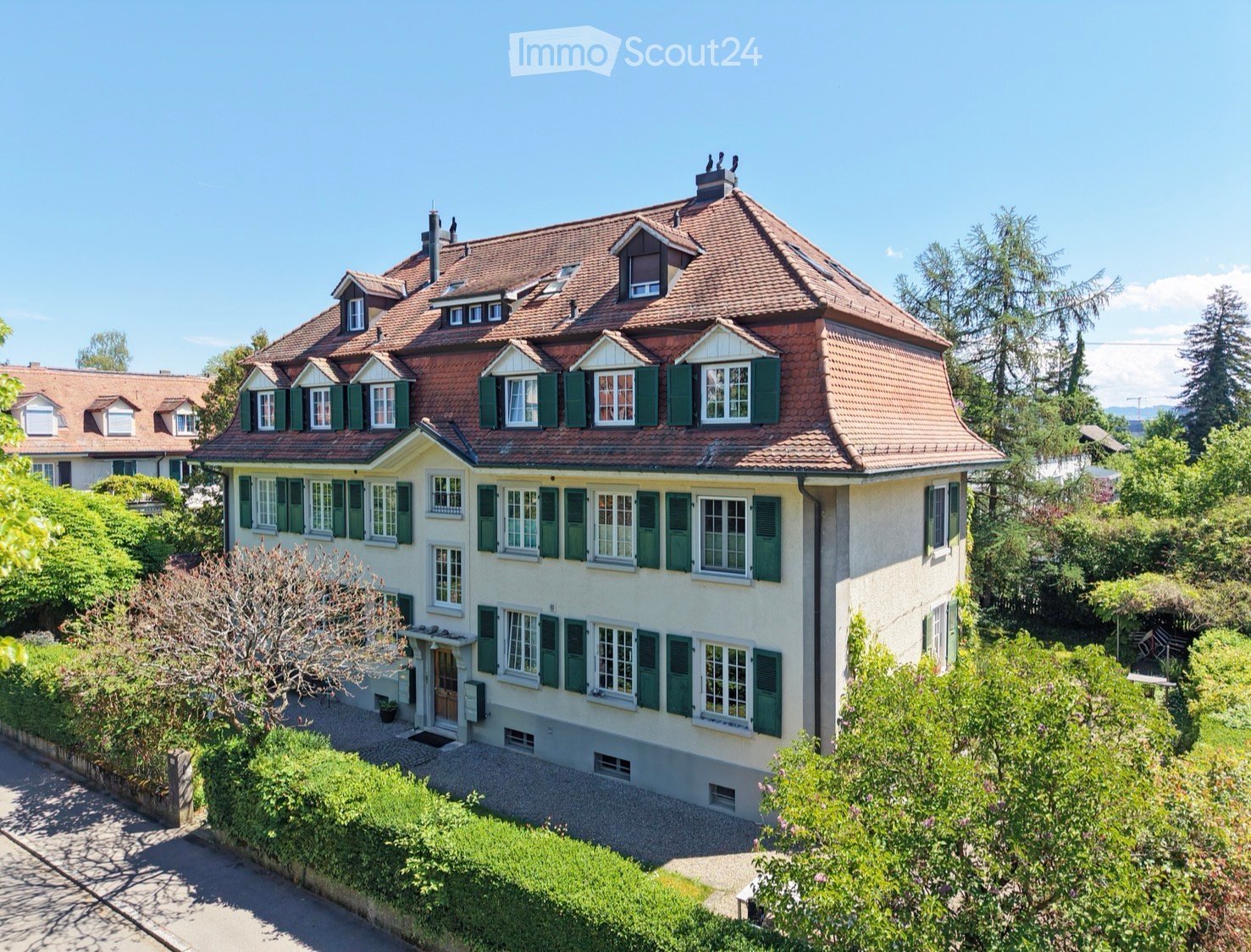 A two-story house with a red-tiled roof, green shutters, and multiple windows, surrounded by lush greenery and trees.