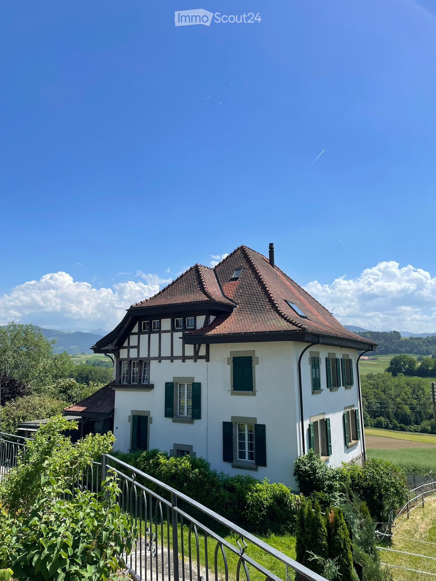 A two-story white house with a red-tiled roof, green shutters, and a balcony overlooking a scenic valley with mountains in the background.
