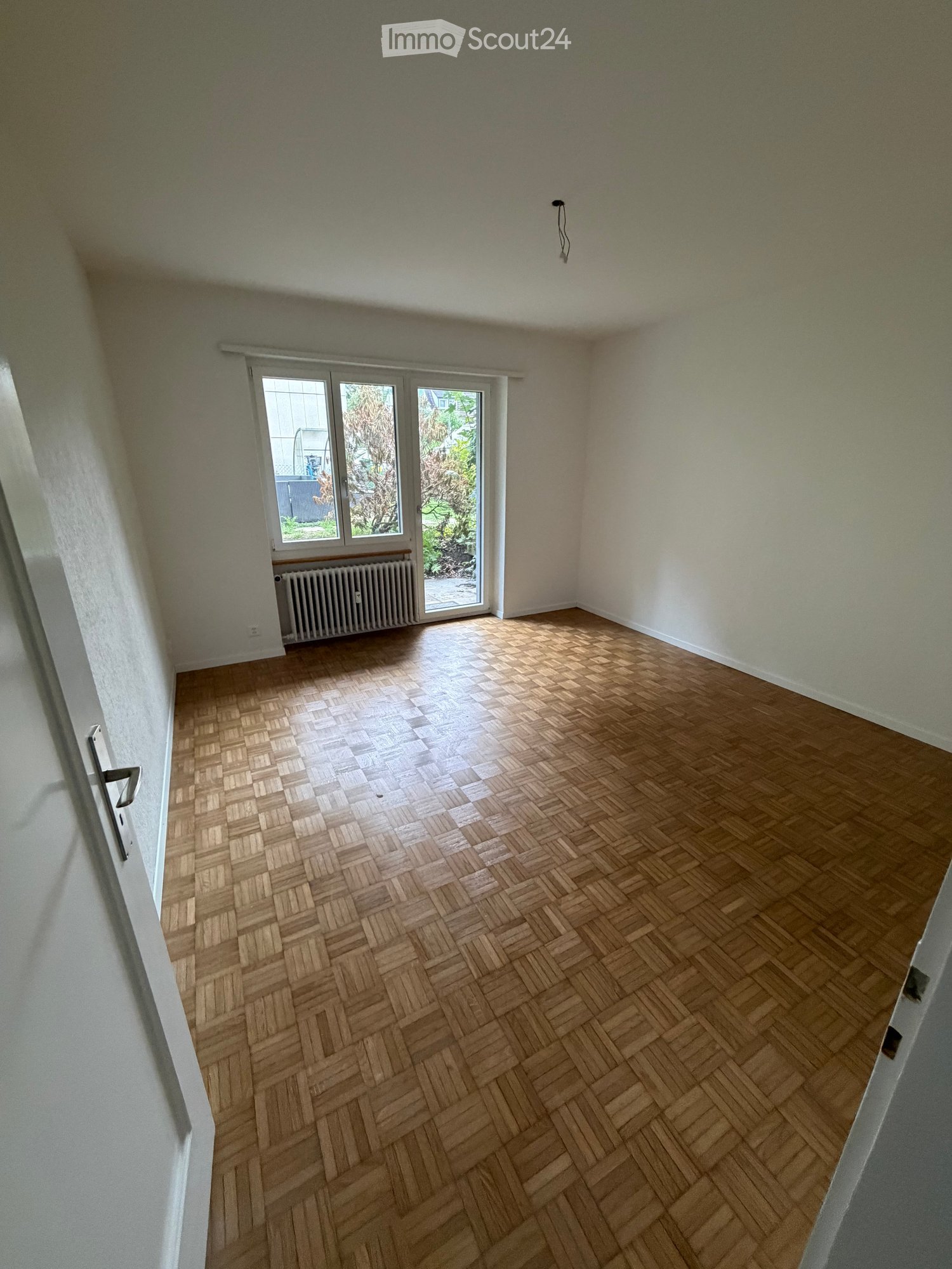 Empty room with wooden herringbone flooring, white walls, and a large window with a radiator below it.