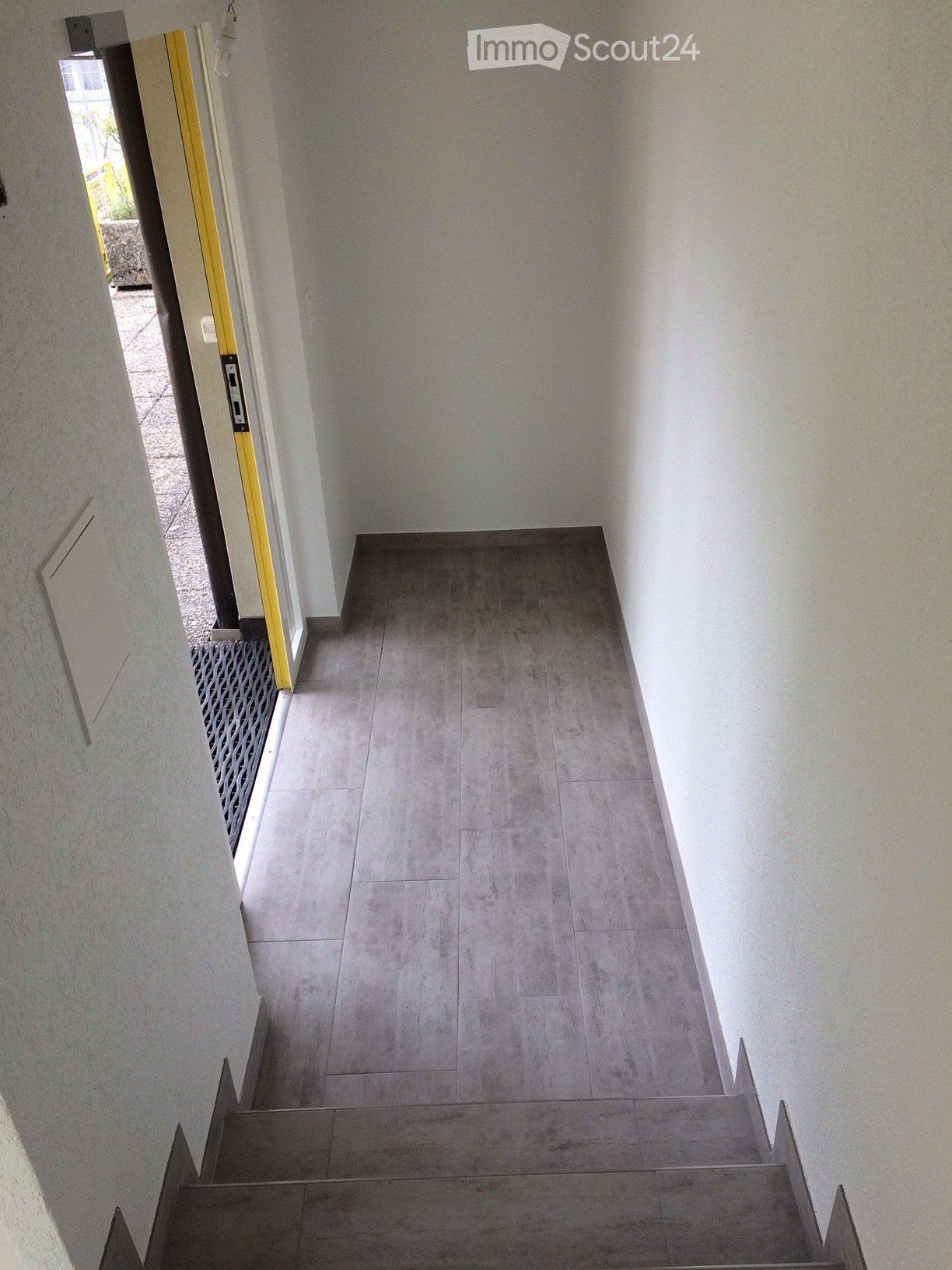 A clean, modern hallway with grey tiled flooring and white walls, featuring a staircase leading upwards and an open doorway to the left.