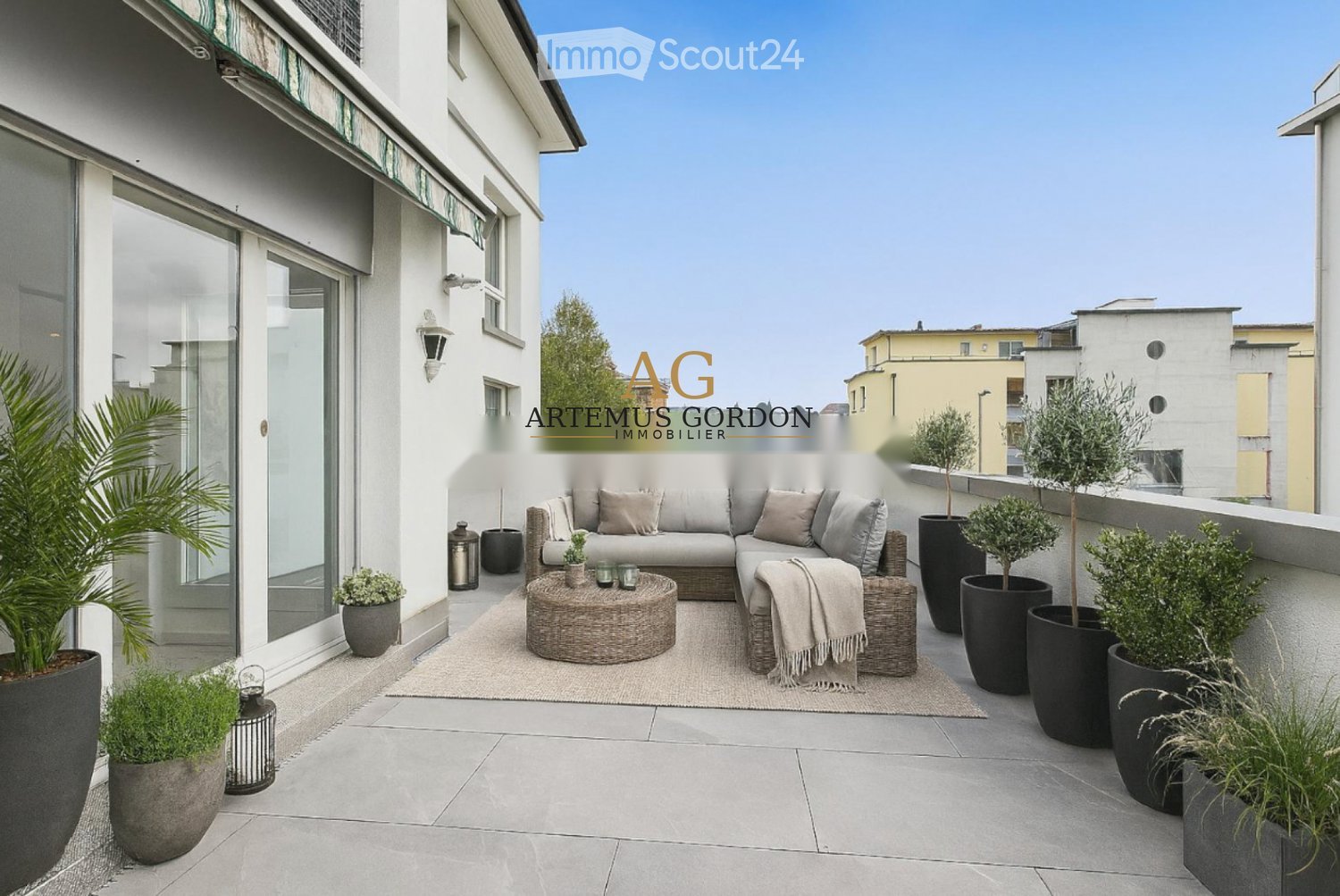 A modern balcony with a sofa, coffee table, and potted plants arranged on a tiled floor.