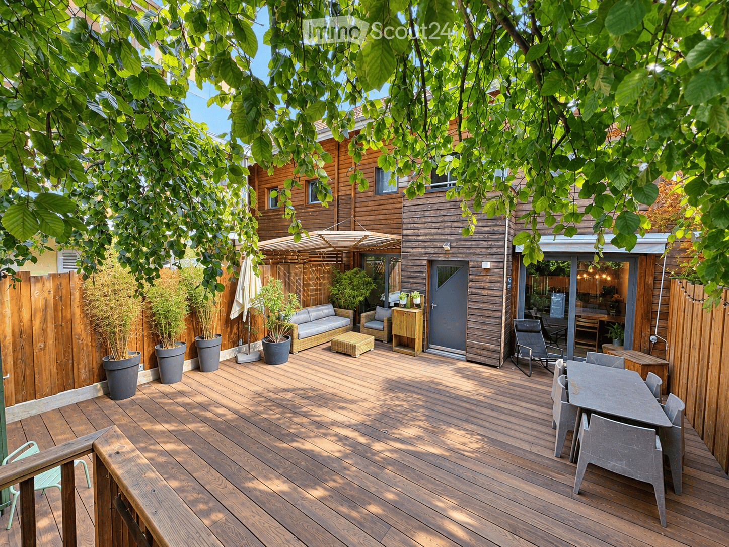 Wooden decked outdoor area with a sofa, coffee table, dining table, chairs, and potted plants, surrounded by wooden fencing and trees.