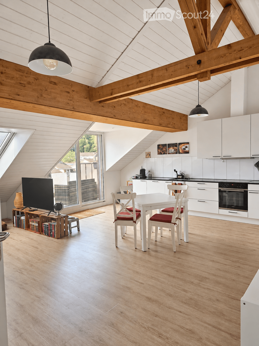 Open-plan living and dining area with exposed wooden beams, white cabinets, a dining table with chairs, a television, and a small bookshelf with books and a vase.