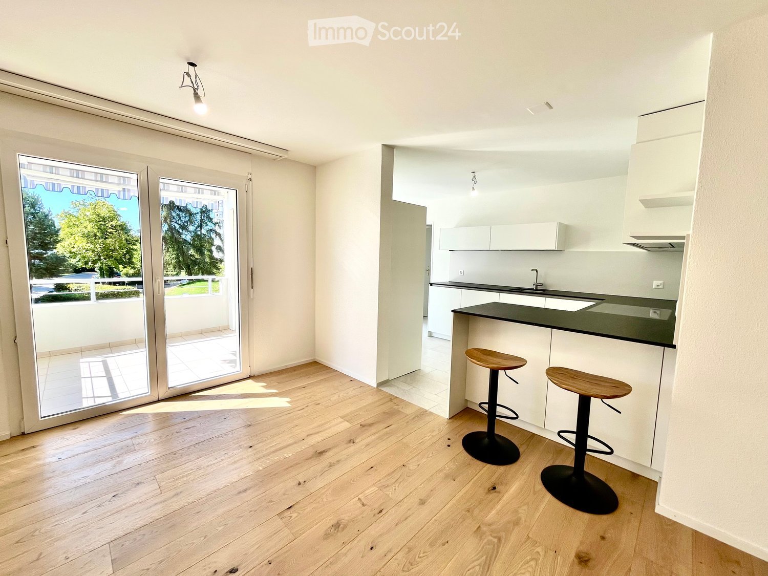 Modern kitchen with light wooden flooring, white cabinets, black countertop, and two wooden stools. Large sliding glass doors lead to a balcony with outdoor view.