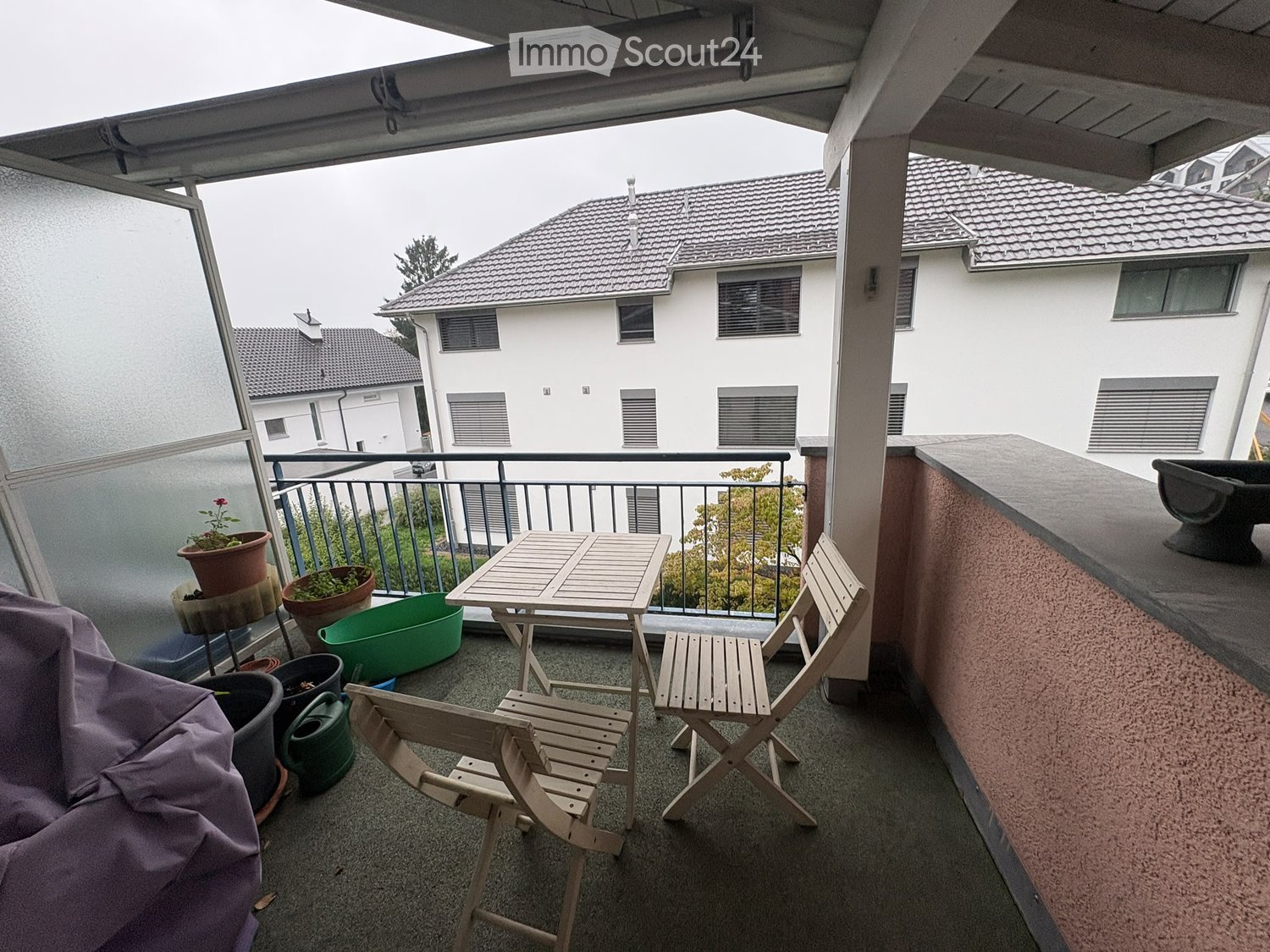 Balcony with a small table, two chairs, several potted plants, and a garden hose. The balcony has a railing and overlooks neighboring buildings.