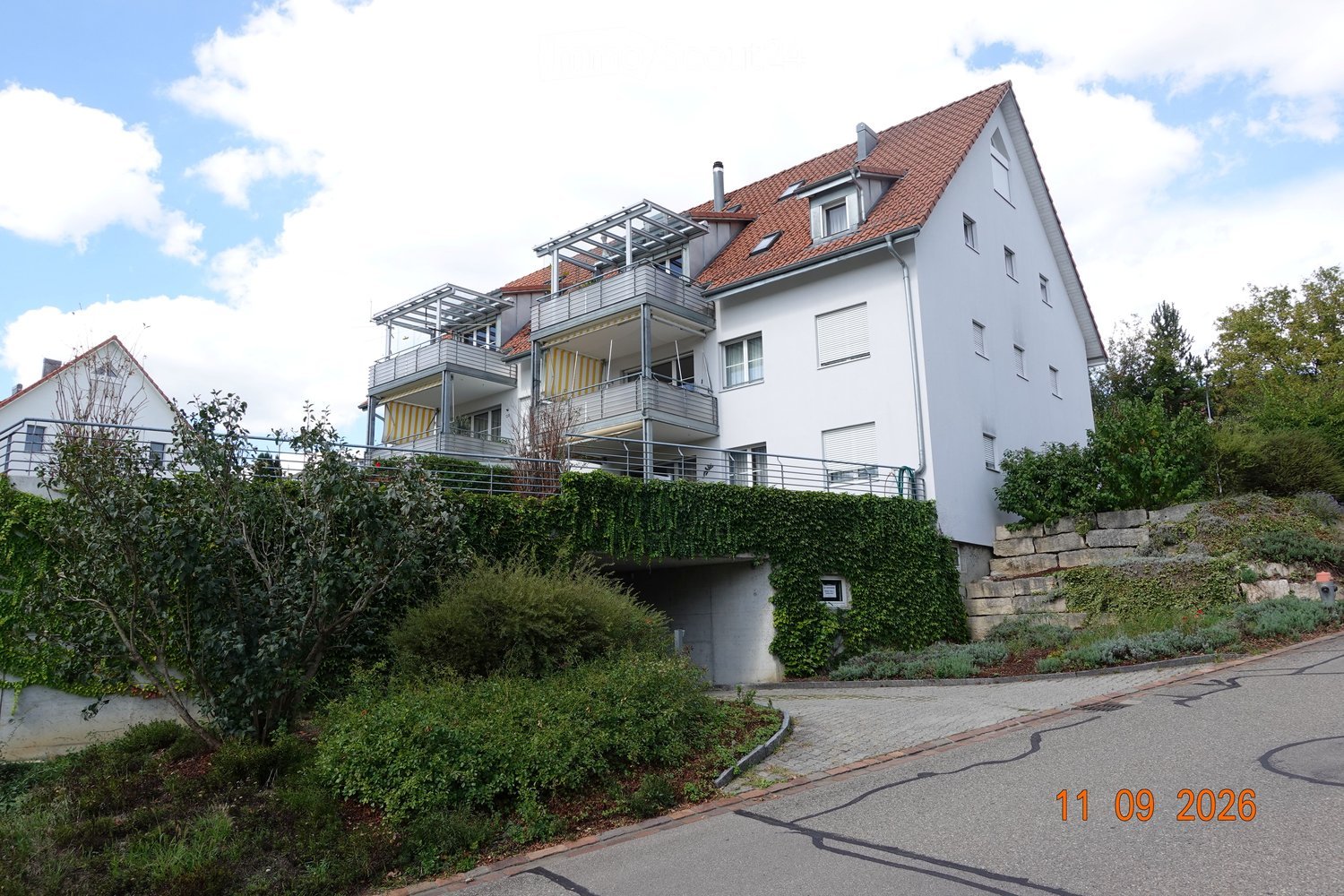Modern two-story house with white exterior, red-tiled roof, multiple windows, balconies, and a well-maintained garden.