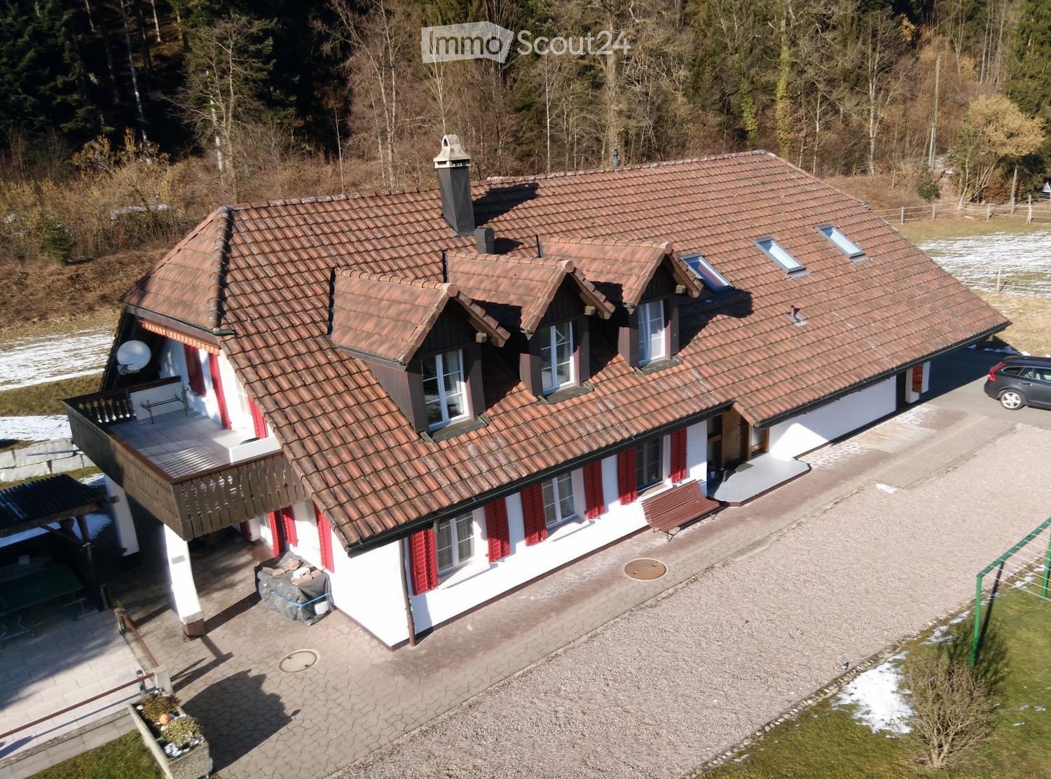 Two-story house with red-tiled roof, white walls, red shutters, balcony, and driveway with parked car.