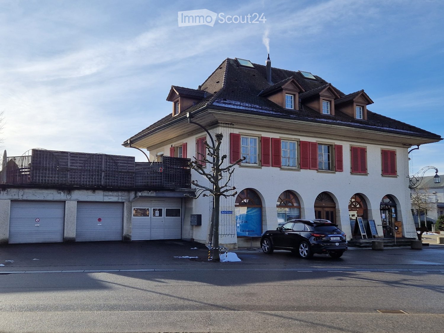 Two story building with red shutters, chimney, garages, a black car parked in front.