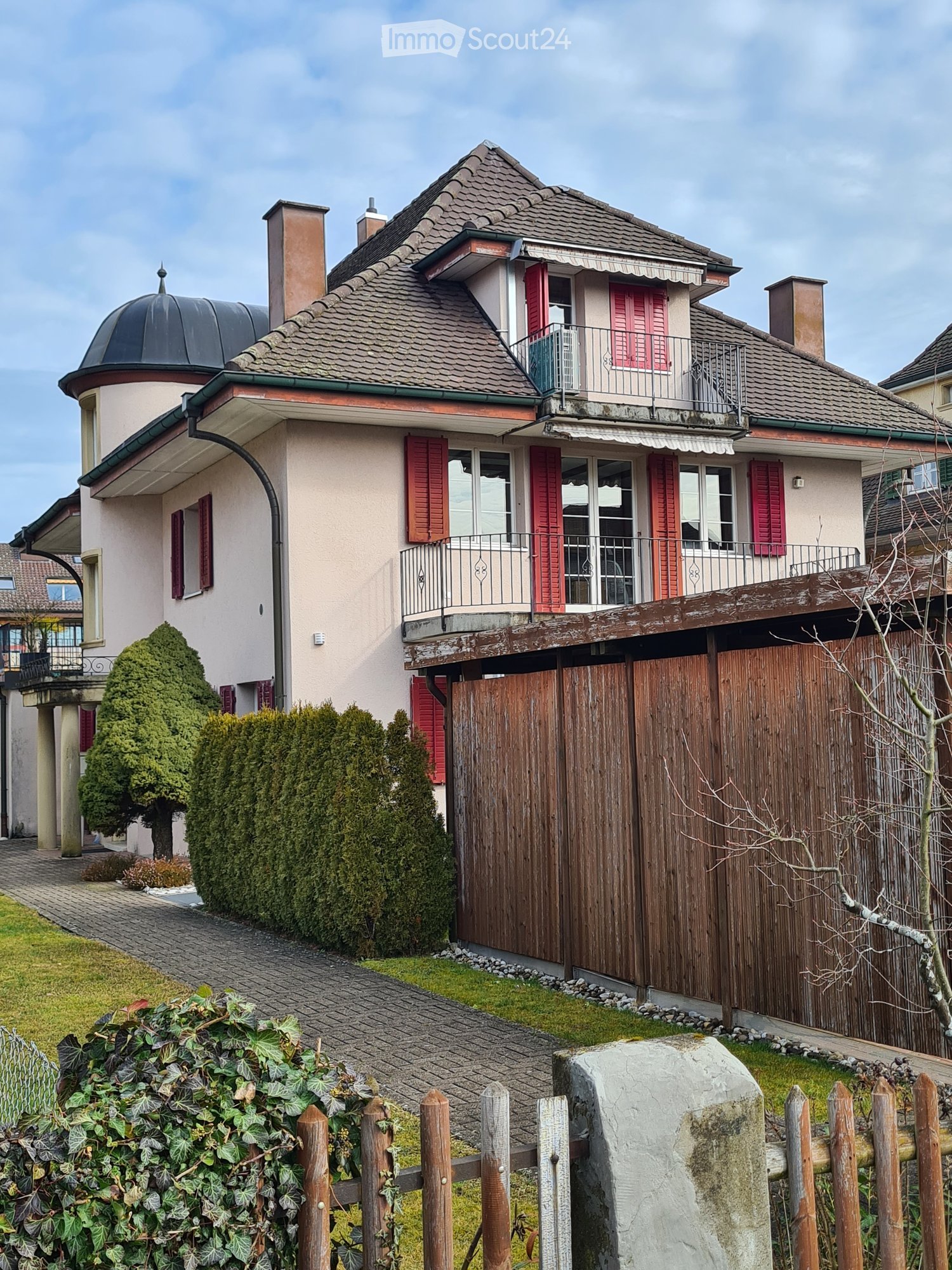Two-story house with red shutters, a balcony, and a wooden gate. The house features a dome-shaped roof section and is surrounded by greenery and a paved pathway.