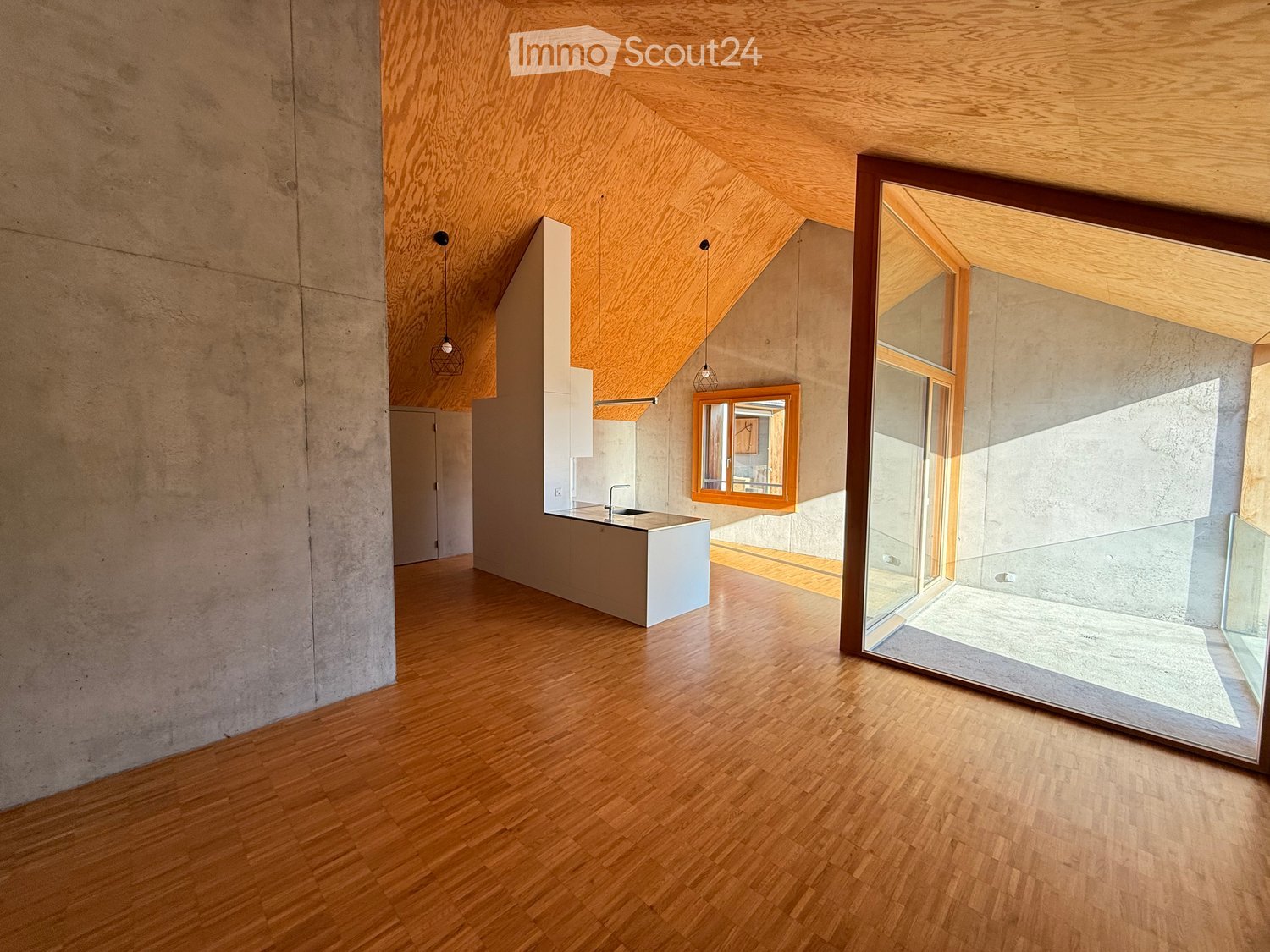 An empty room with wooden flooring, concrete walls, and a large window. The room features a minimalist design with exposed wooden beams on the ceiling and a small kitchen area visible in the background.