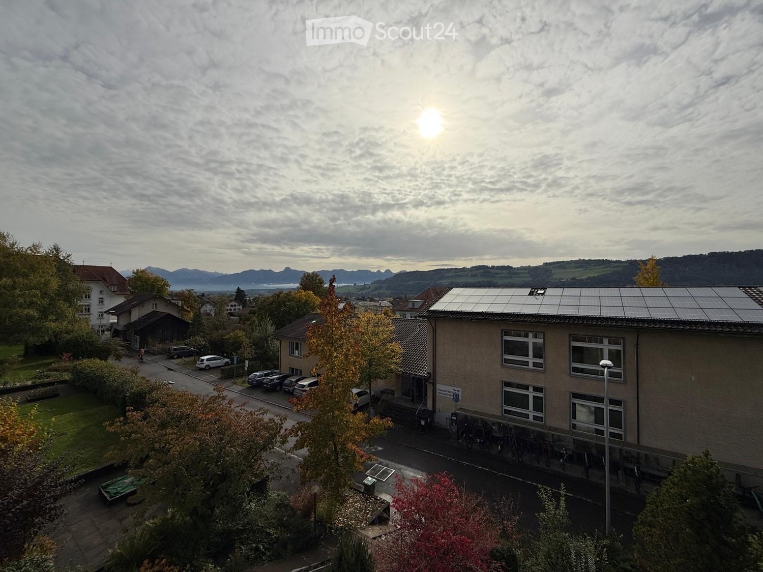 A multi-story building with solar panels on the roof, surrounded by trees and parked cars, with a scenic mountain view in the background.