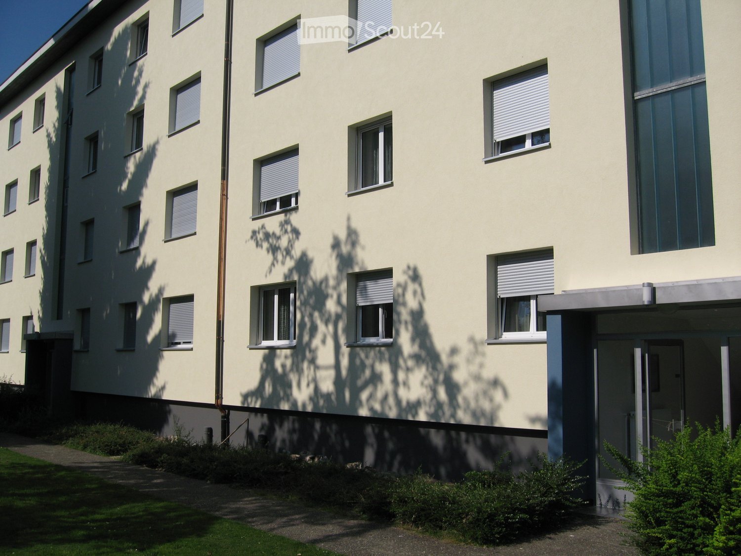 Four-story building with light yellow facade, multiple windows with blinds, and a small entrance area with greenery.