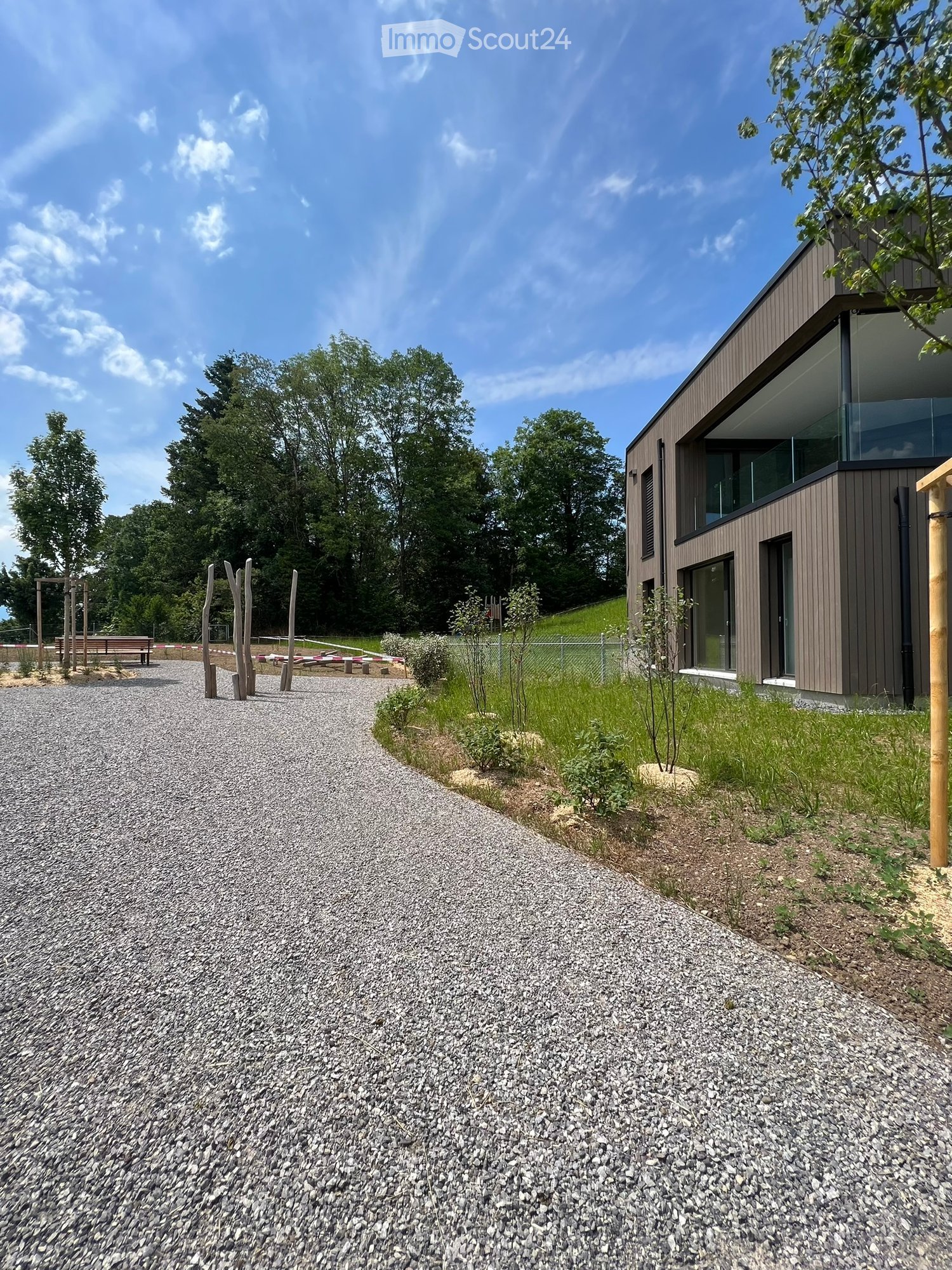 Modern two-story house with dark brown siding and large windows, surrounded by a gravel pathway and greenery.