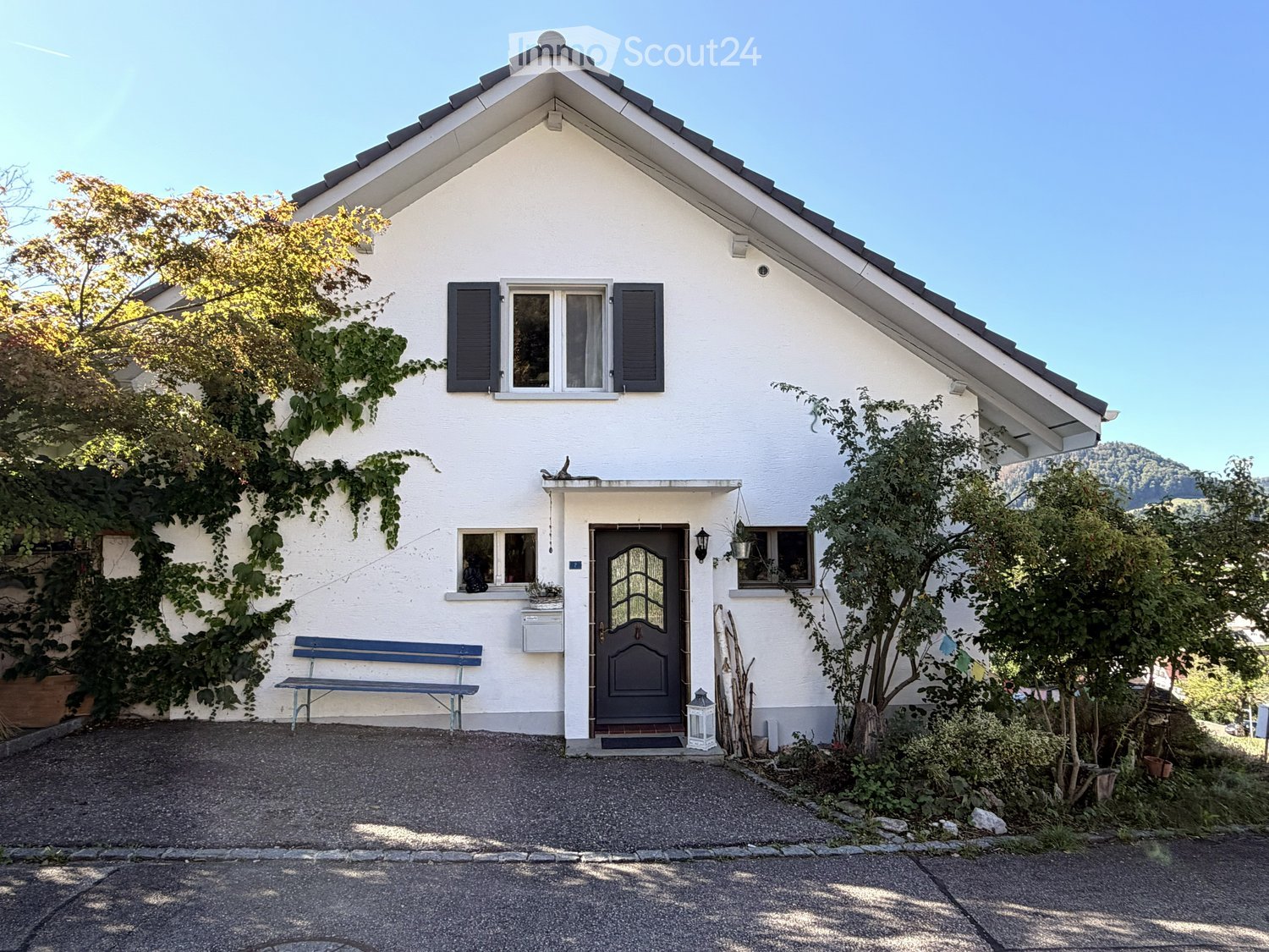 Single-story white house with dark shutters, a front door with glass panels, and a small bench near the entrance. The house is surrounded by greenery and has a gravel driveway.