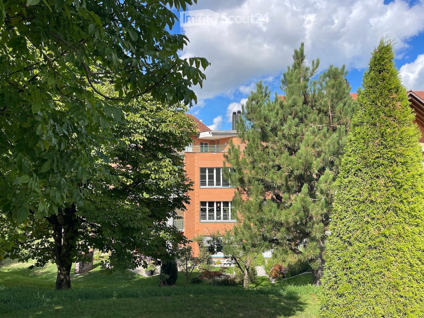 A two-story house with orange brick walls, white-framed windows, and a red-tiled roof, surrounded by lush greenery and trees.