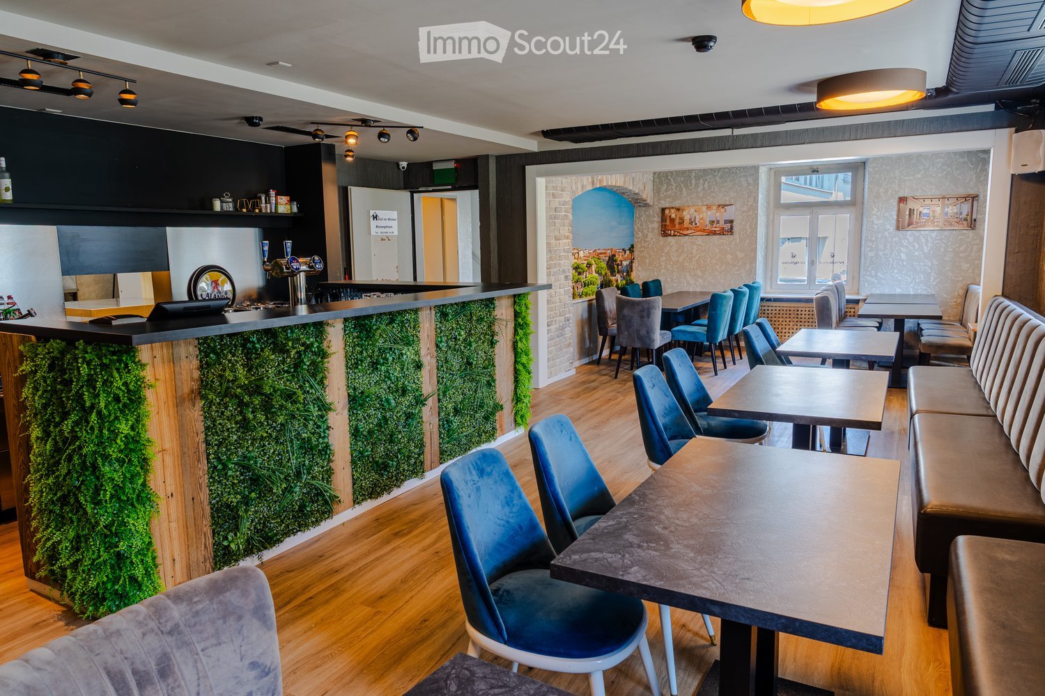 Modern restaurant interior with wooden flooring, blue chairs, tables, a bar counter with a green moss wall, and a seating area with a sofa and bench.