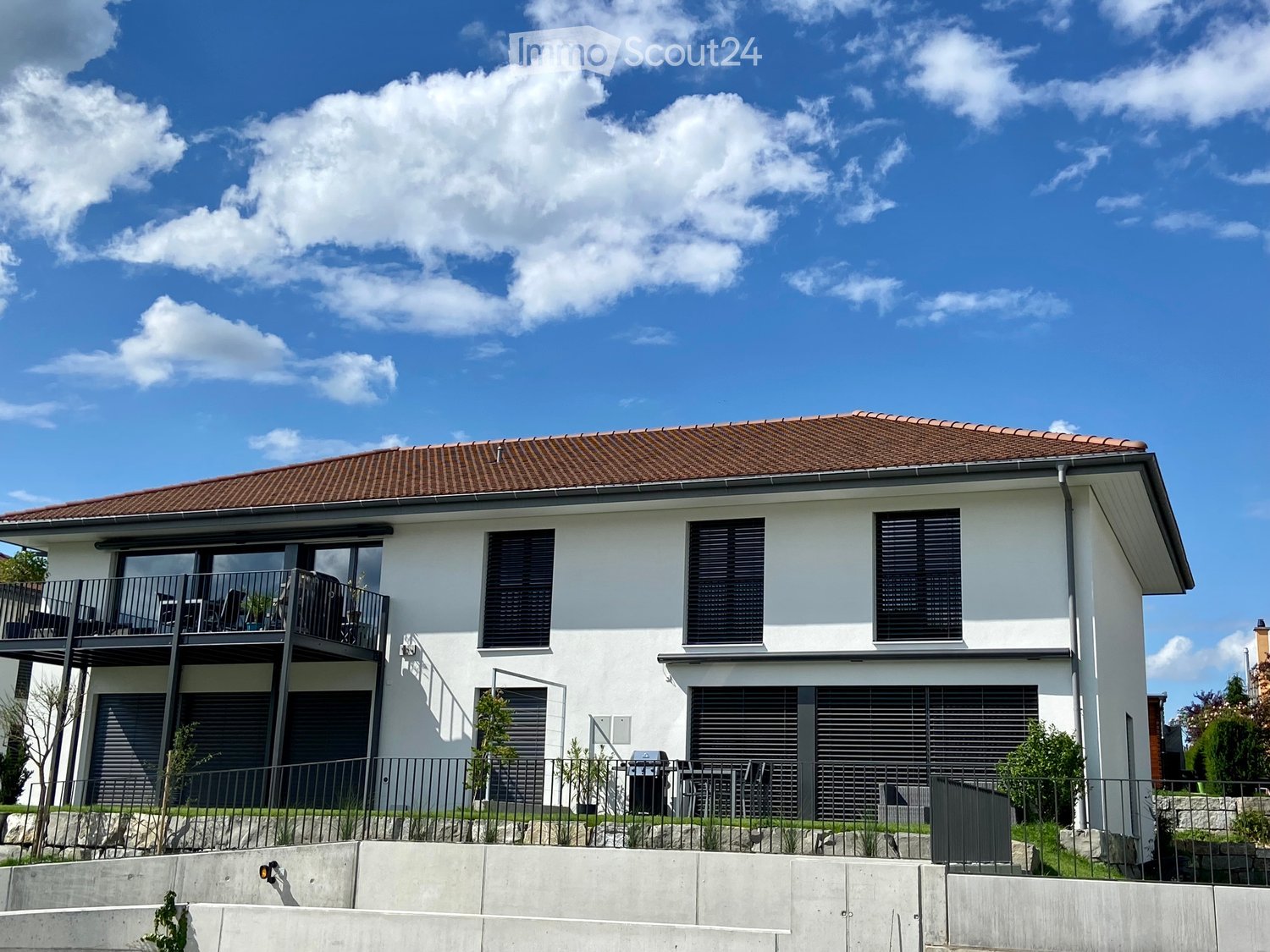 Modern two-story house with white exterior, brown tiled roof, large windows, and a balcony on the upper level. Features include a garage, raised ground floor, and outdoor seating area.
