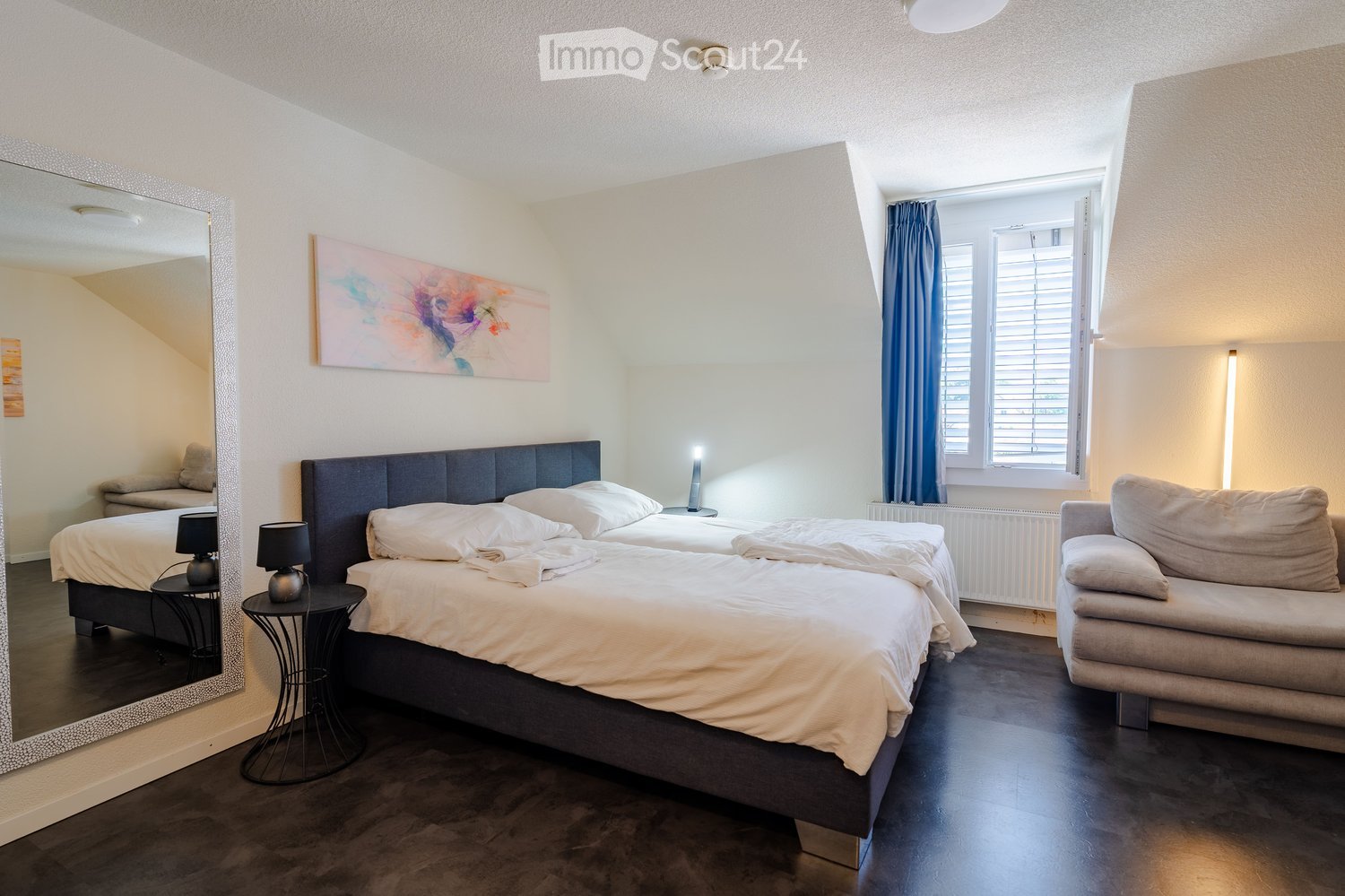 Cozy bedroom with two single beds, a sofa, small side tables with lamps, a large mirror, and a colorful painting on the wall. The room features dark wooden flooring and large windows with blue curtains.
