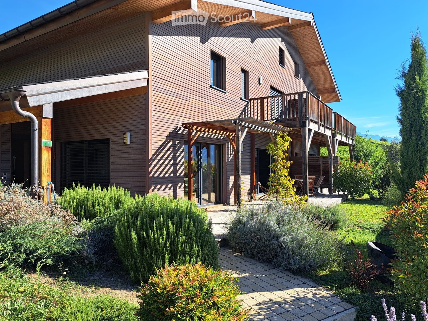 Two-story wooden house with a balcony, covered patio, and well-maintained garden with shrubs and a lawn.