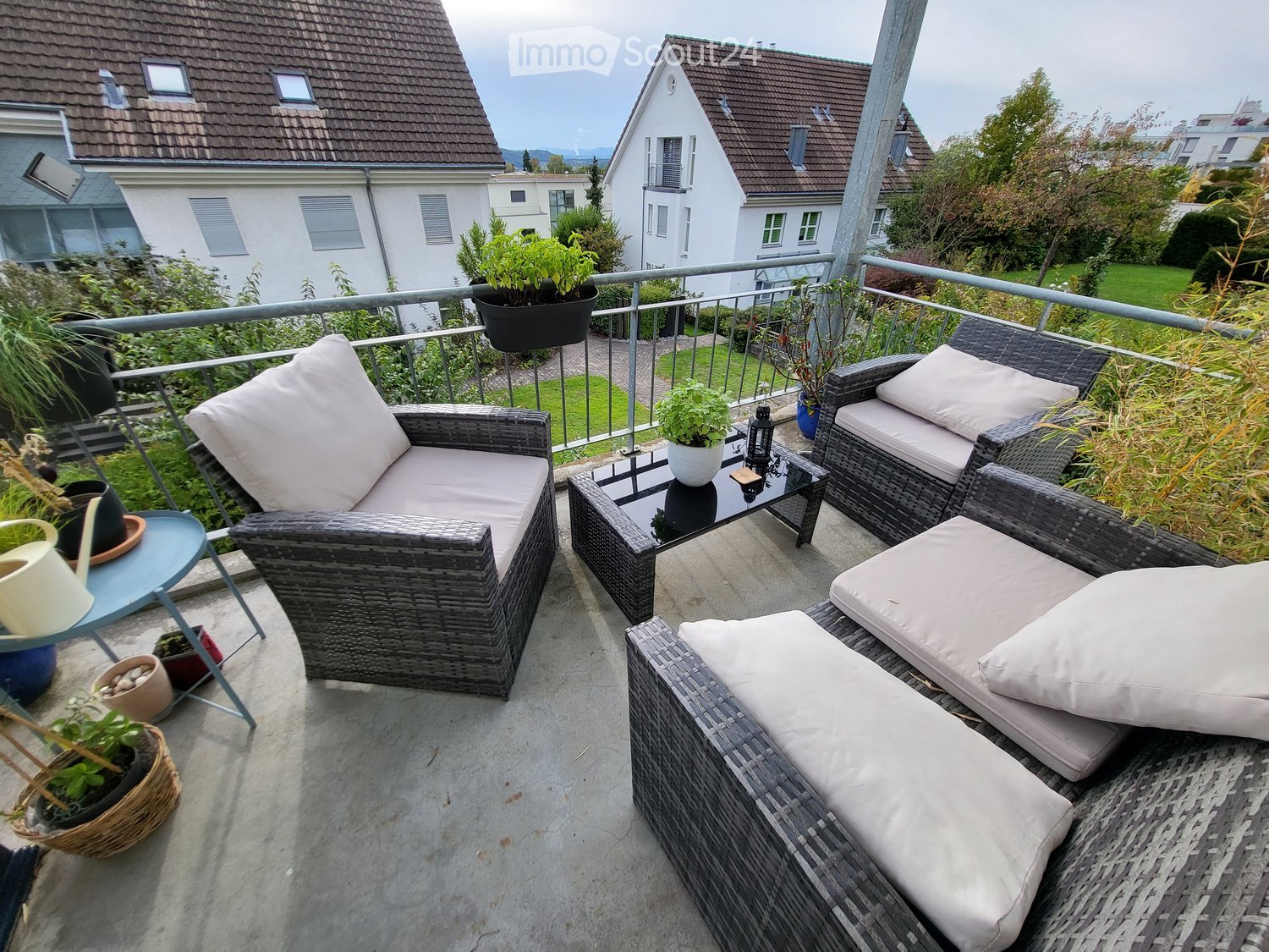 A well-furnished balcony with wicker chairs, a sofa, a coffee table, and several potted plants, overlooking a residential area.