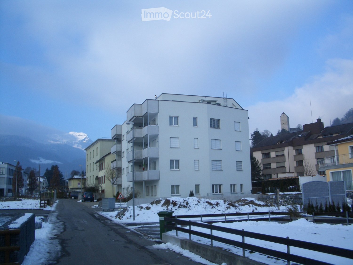 Multi-story residential building with balconies and windows, surrounded by other buildings and a snow-covered landscape.
