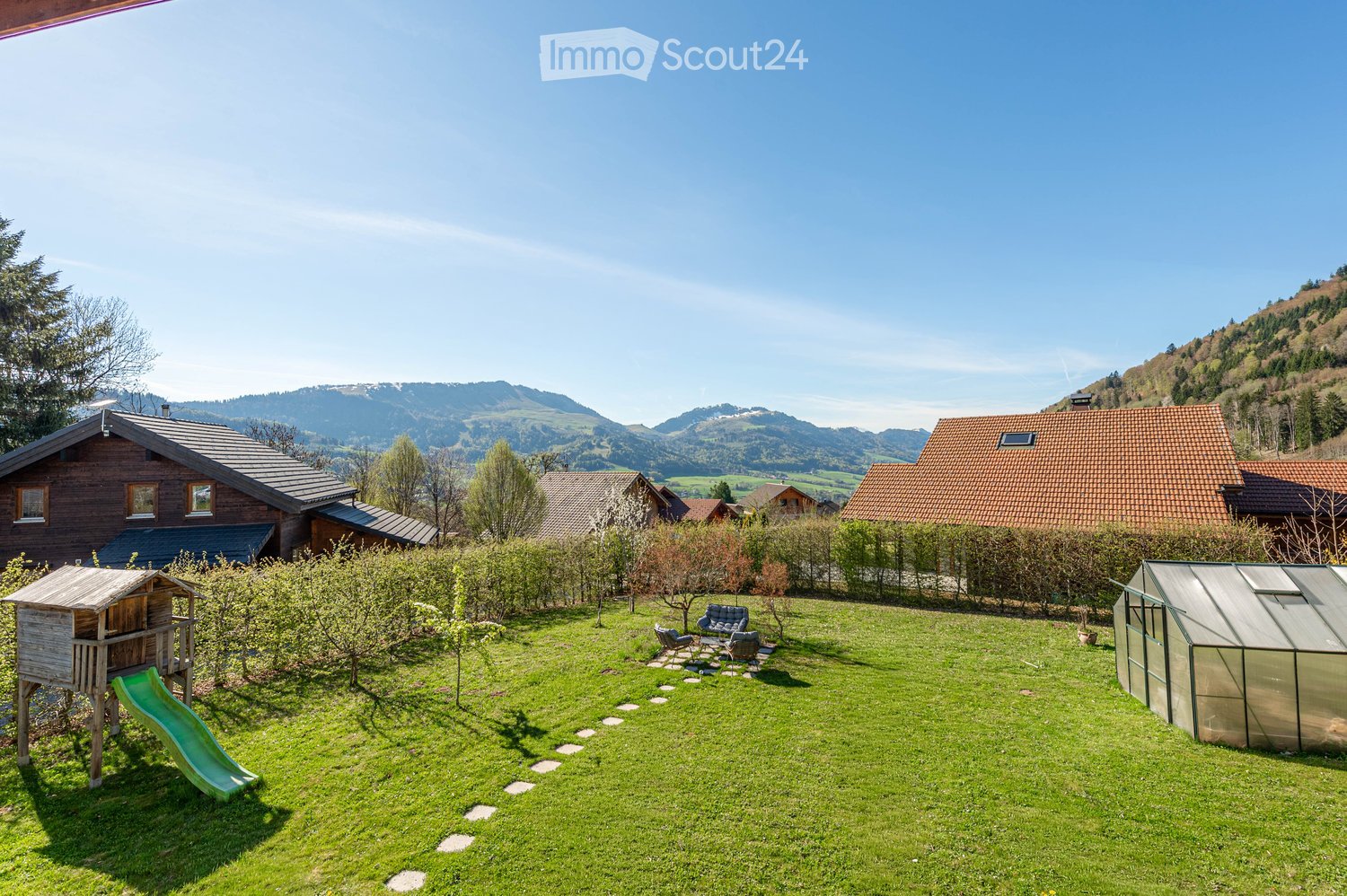 A spacious garden with a wooden playhouse, green slide, patio furniture, greenhouse, and surrounding trees and mountains.