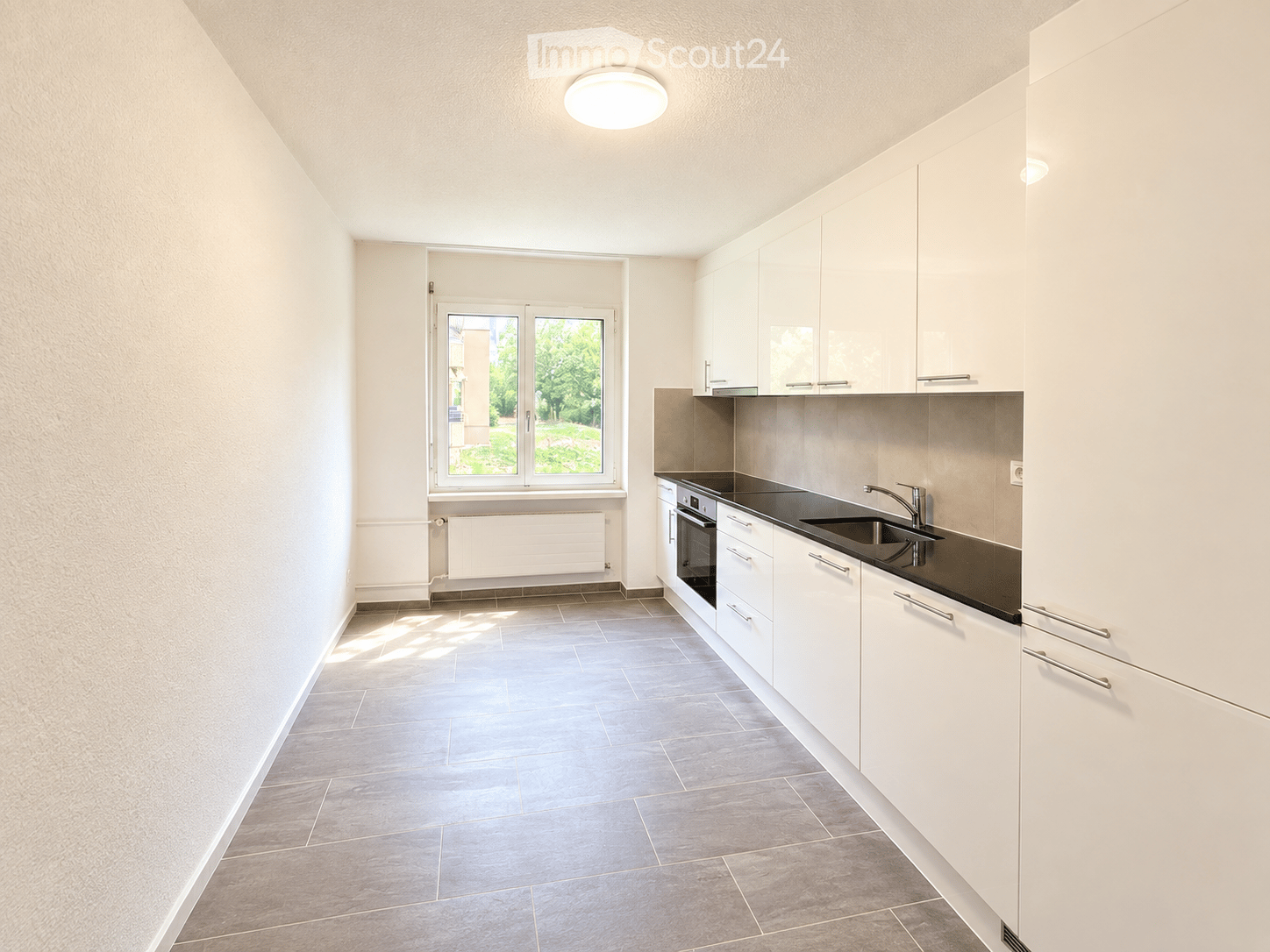 Modern kitchen with white cabinets, dark countertops, and a built-in oven. Features a large window with a view of greenery outside.