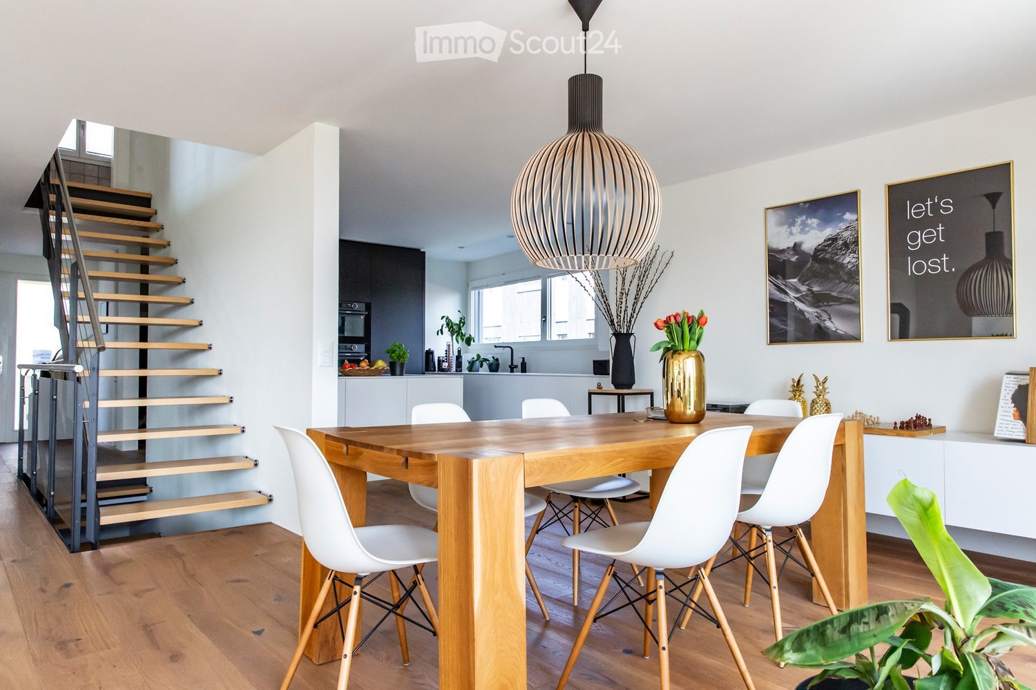 Modern dining area with a wooden table, white chairs, a large pendant light, and a staircase leading to an upper level. The space includes a kitchen area with an oven, sink, and various decorative items such as vases, picture frames, and plants.