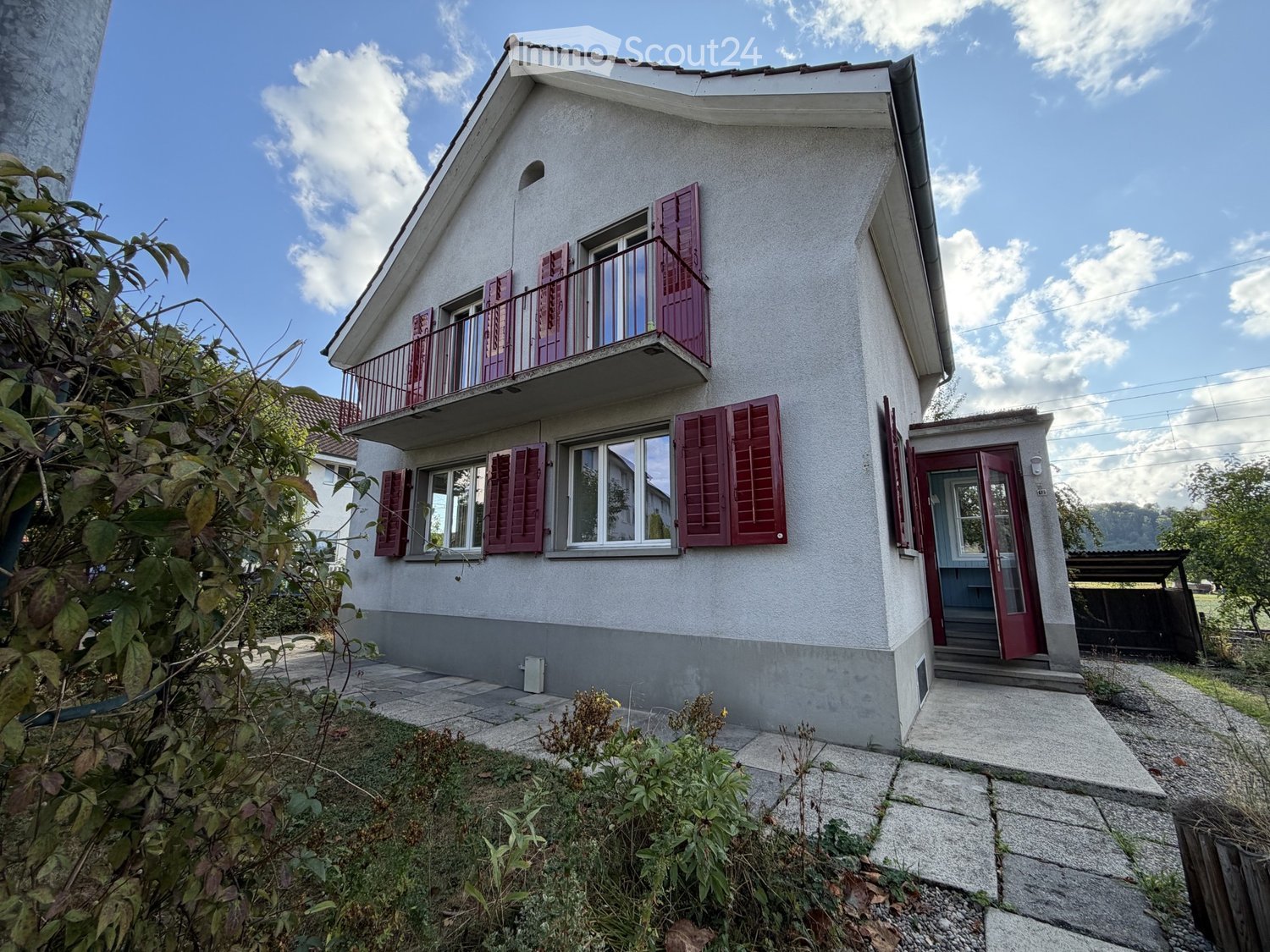 Two-story house with white exterior, red shutters, and a balcony on the upper floor. The house features a small garden area in front with a stone pathway leading to the entrance.